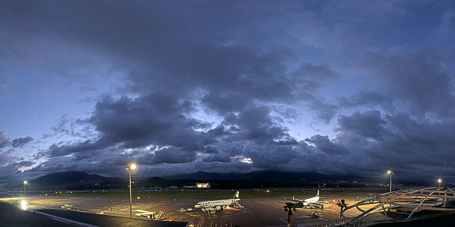 Under a dark, cloudy twilight sky, two airplanes are parked on an airport tarmac, surrounded by airport buildings and lights.