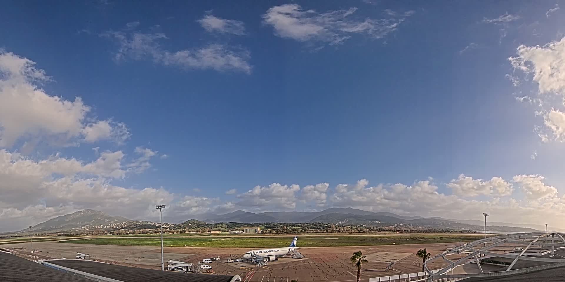 A mostly sunny day with scattered clouds shows an airplane on the tarmac of an airport, with mountains visible in the distance.