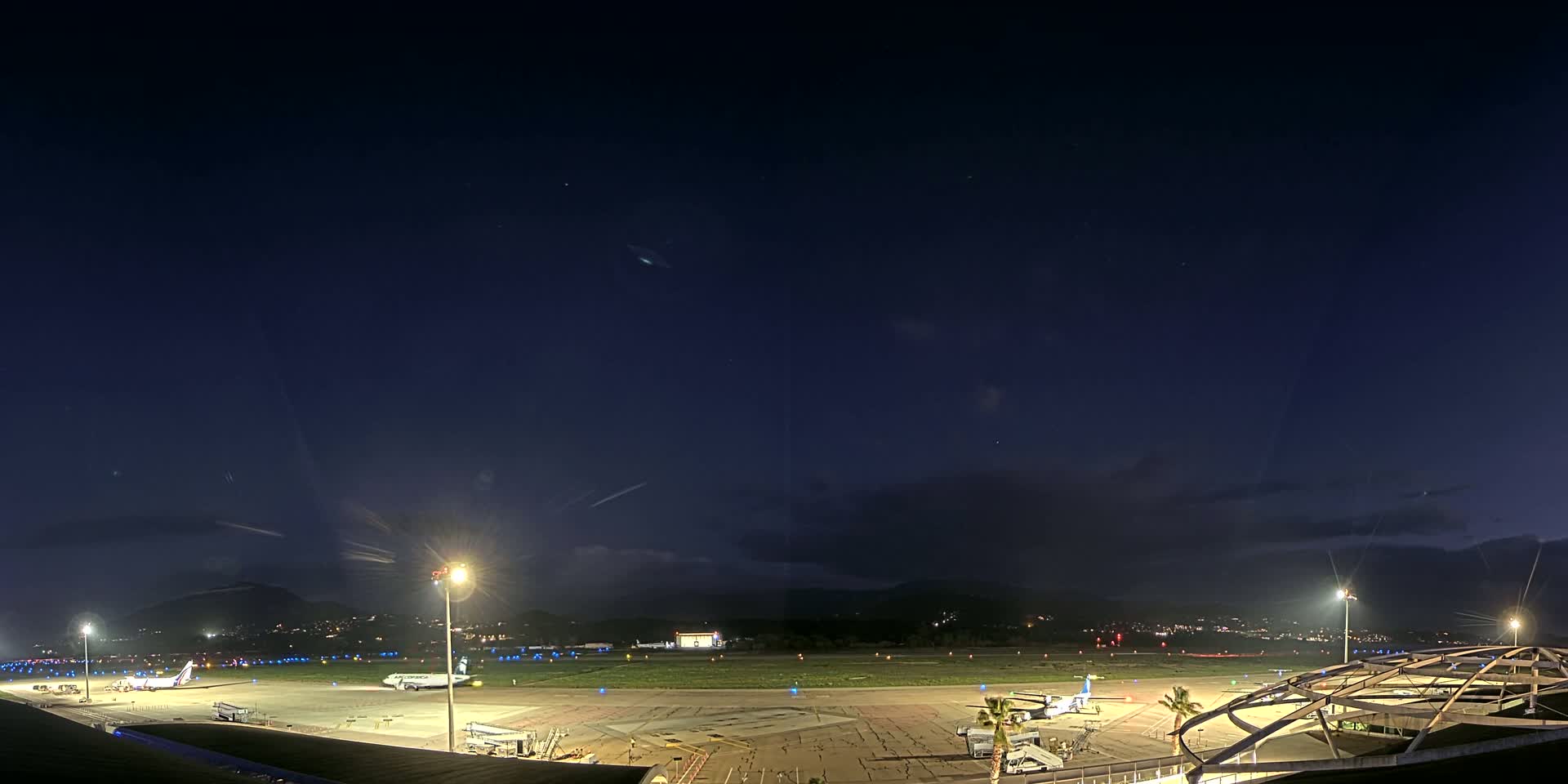 A nighttime view of an airport tarmac with several airplanes, ground support equipment, and illuminated buildings under a mostly clear, dark blue sky.
