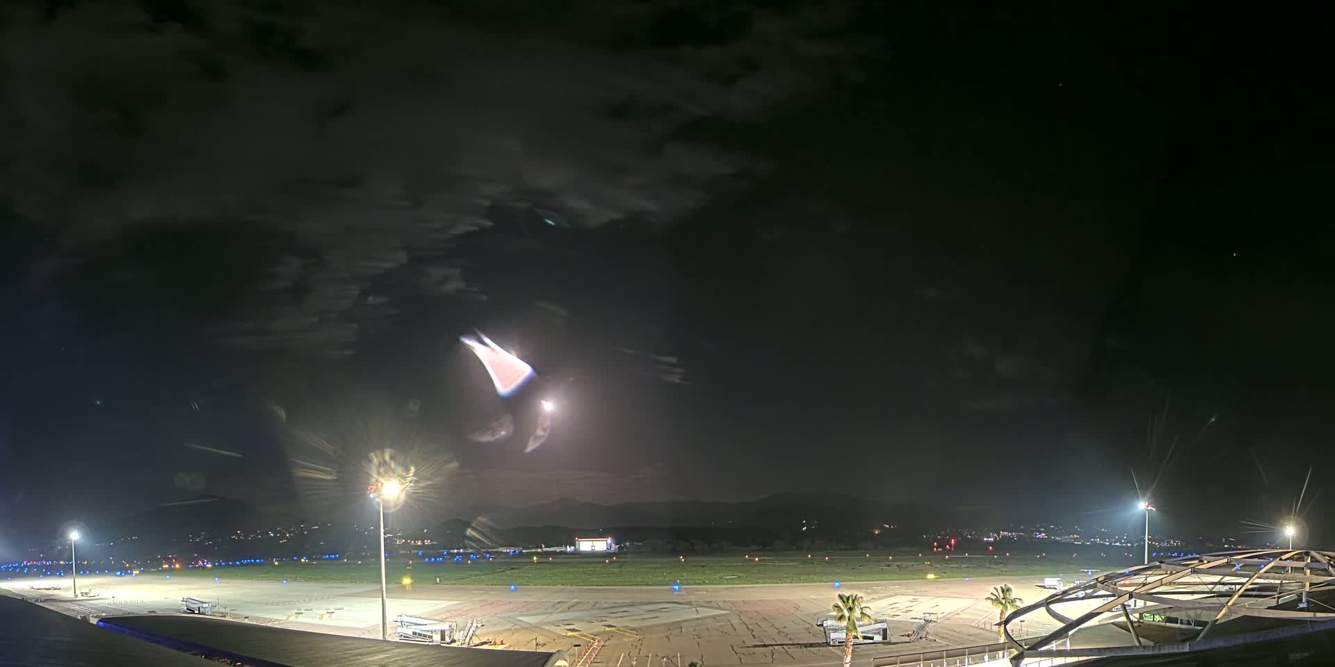 A nighttime view of an airport tarmac with planes parked, illuminated by bright lights under a partly cloudy sky.