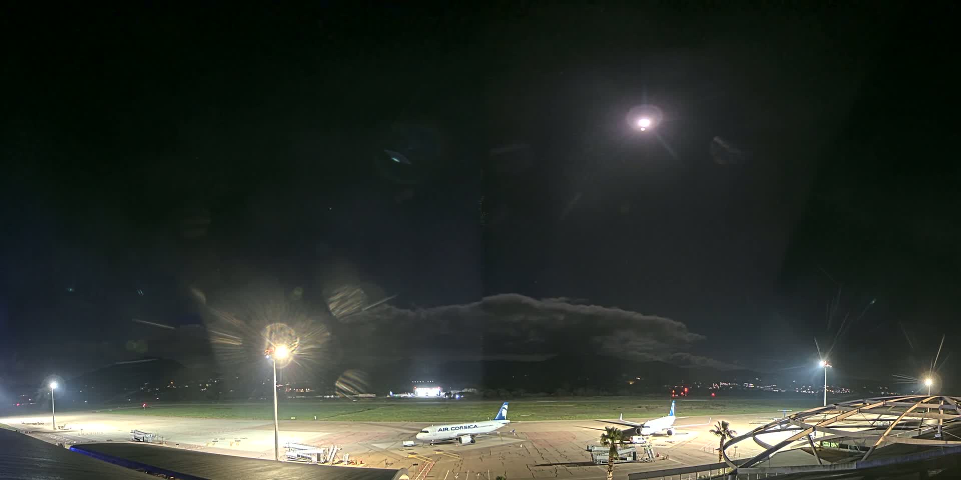 A nighttime view of an airport tarmac with two airplanes parked, illuminated by bright lights under a mostly clear, dark sky with a bright moon.