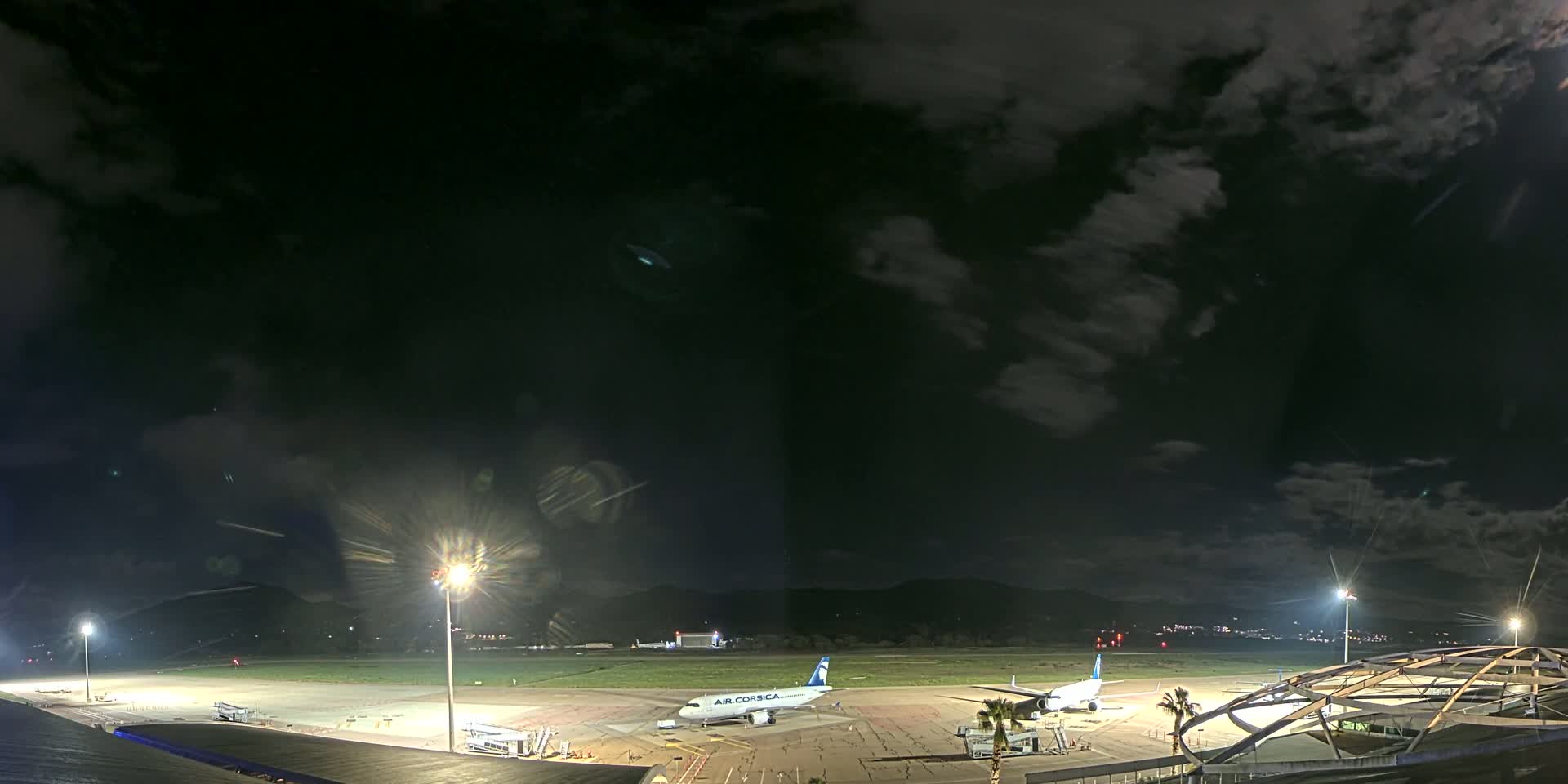 A nighttime view of an airport tarmac with two airplanes parked, under a partly cloudy sky.