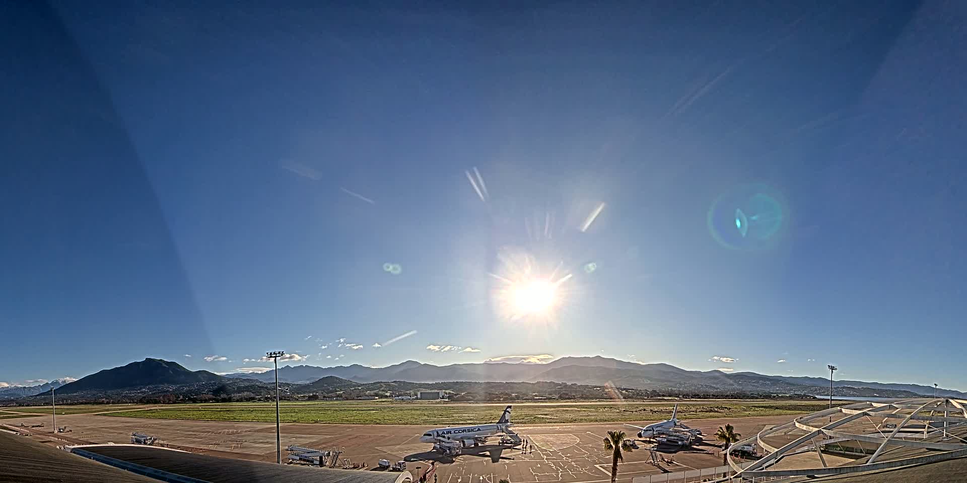 A sunny day at an airport shows several airplanes parked on the tarmac with mountains in the background.