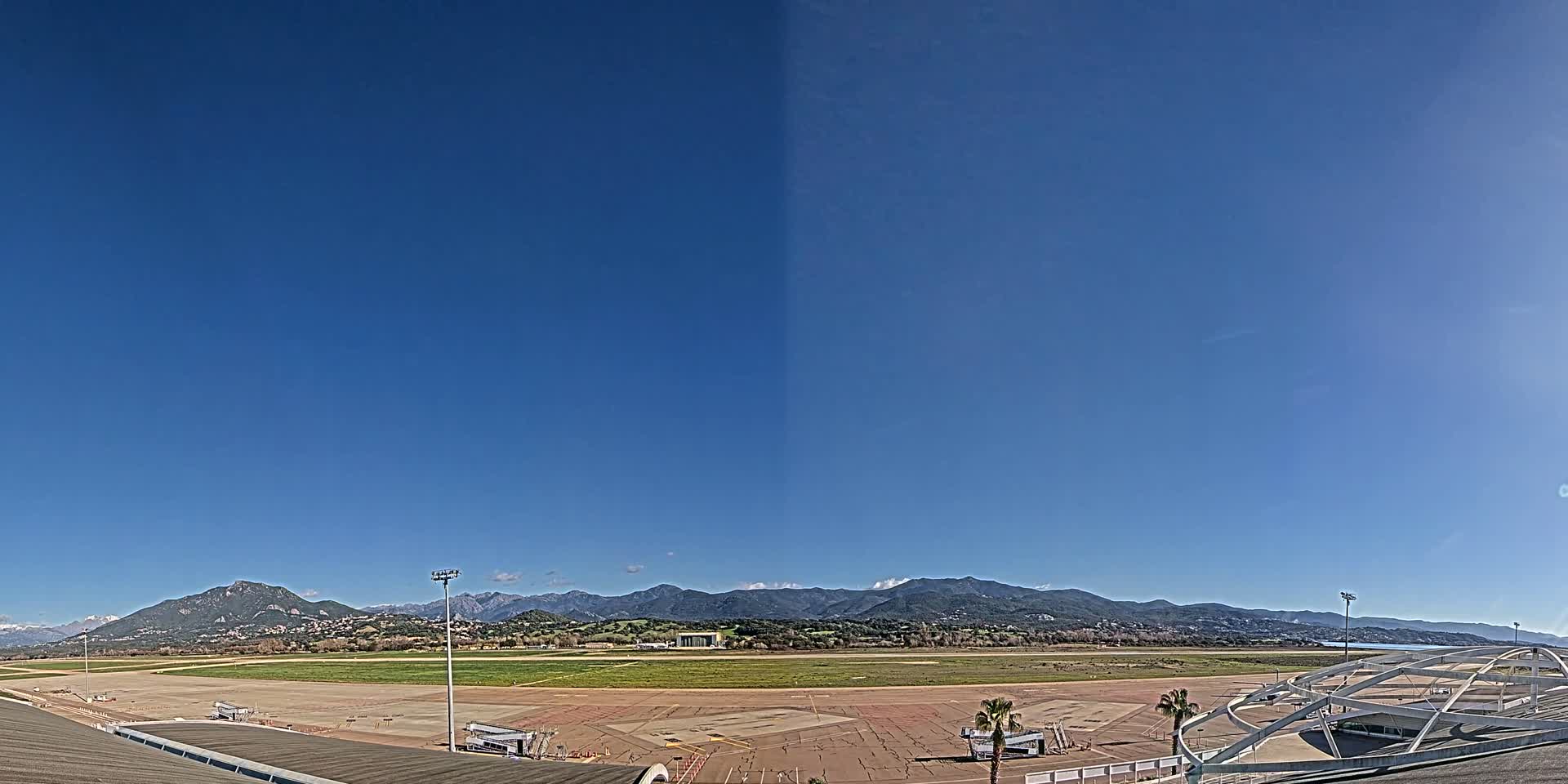 A clear, sunny day reveals a wide expanse of airport tarmac and runway, backed by a range of distant mountains.