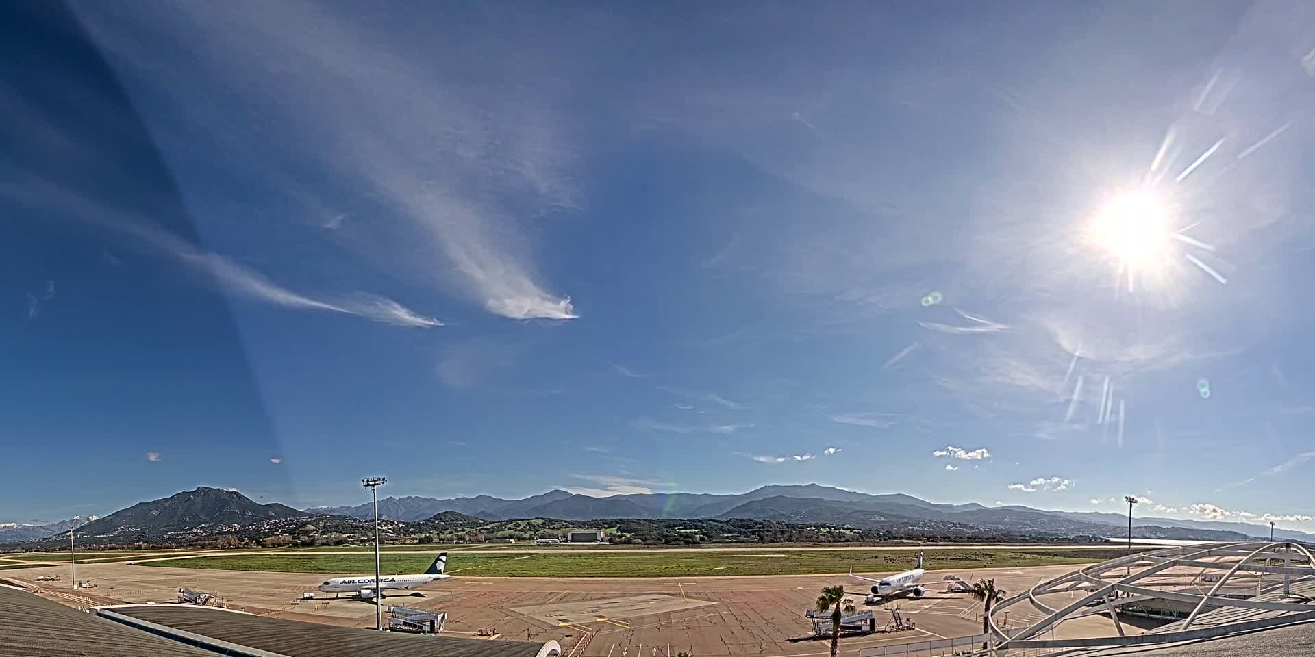 A sunny day at an airport shows two airplanes parked on the tarmac, with mountains visible in the distance under a mostly clear blue sky with wispy clouds.