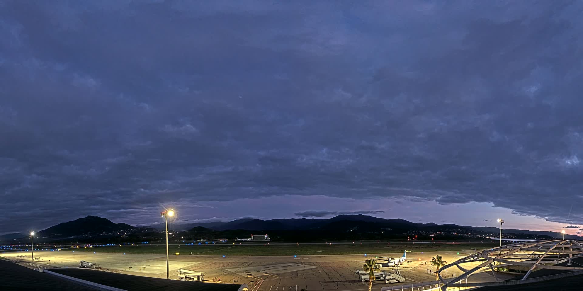 An airport tarmac at twilight under a mostly cloudy sky, with mountains visible in the distance.