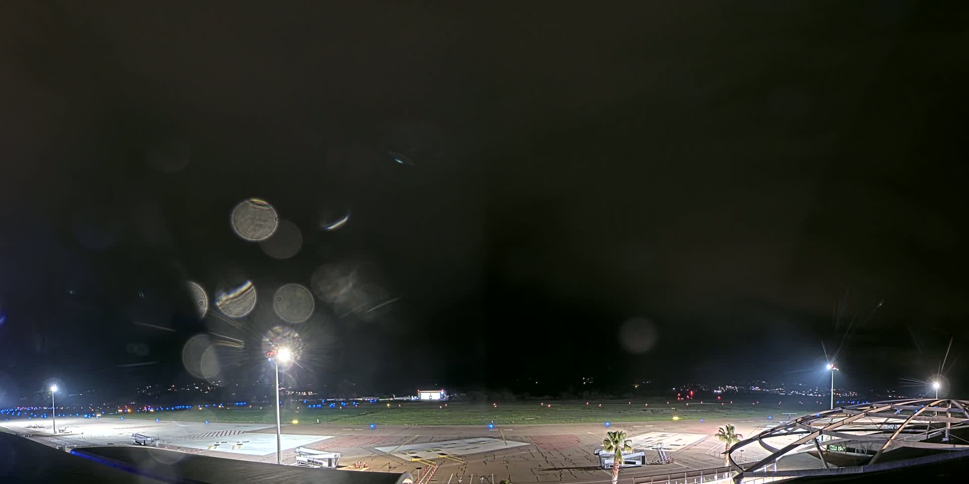 A nighttime view of an airport tarmac, with rain obscuring the view and lights illuminating the runway and surrounding area.