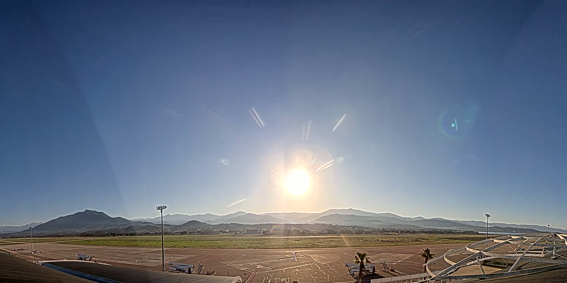 A bright sun shines over an airport tarmac and runway, with mountains visible in the distance under a clear blue sky.