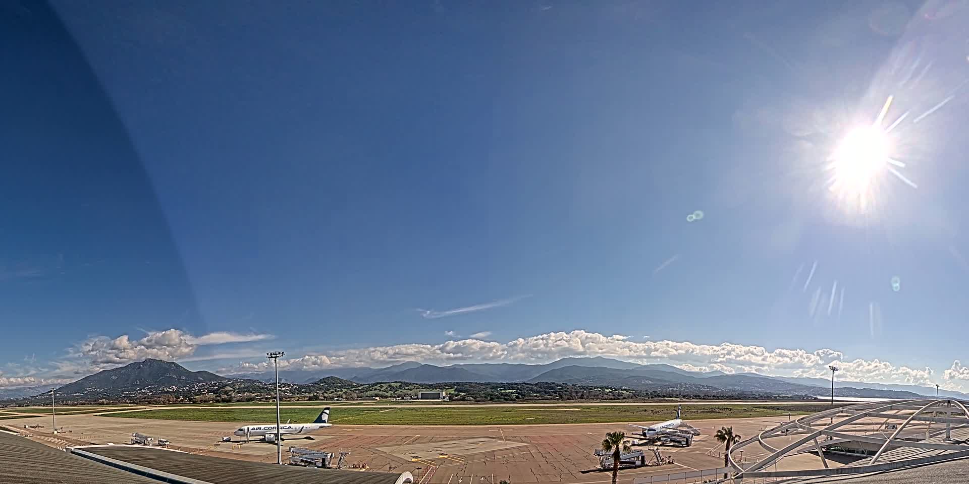 A sunny day at an airport with a few planes on the tarmac, mountains in the background, and partly cloudy skies.
