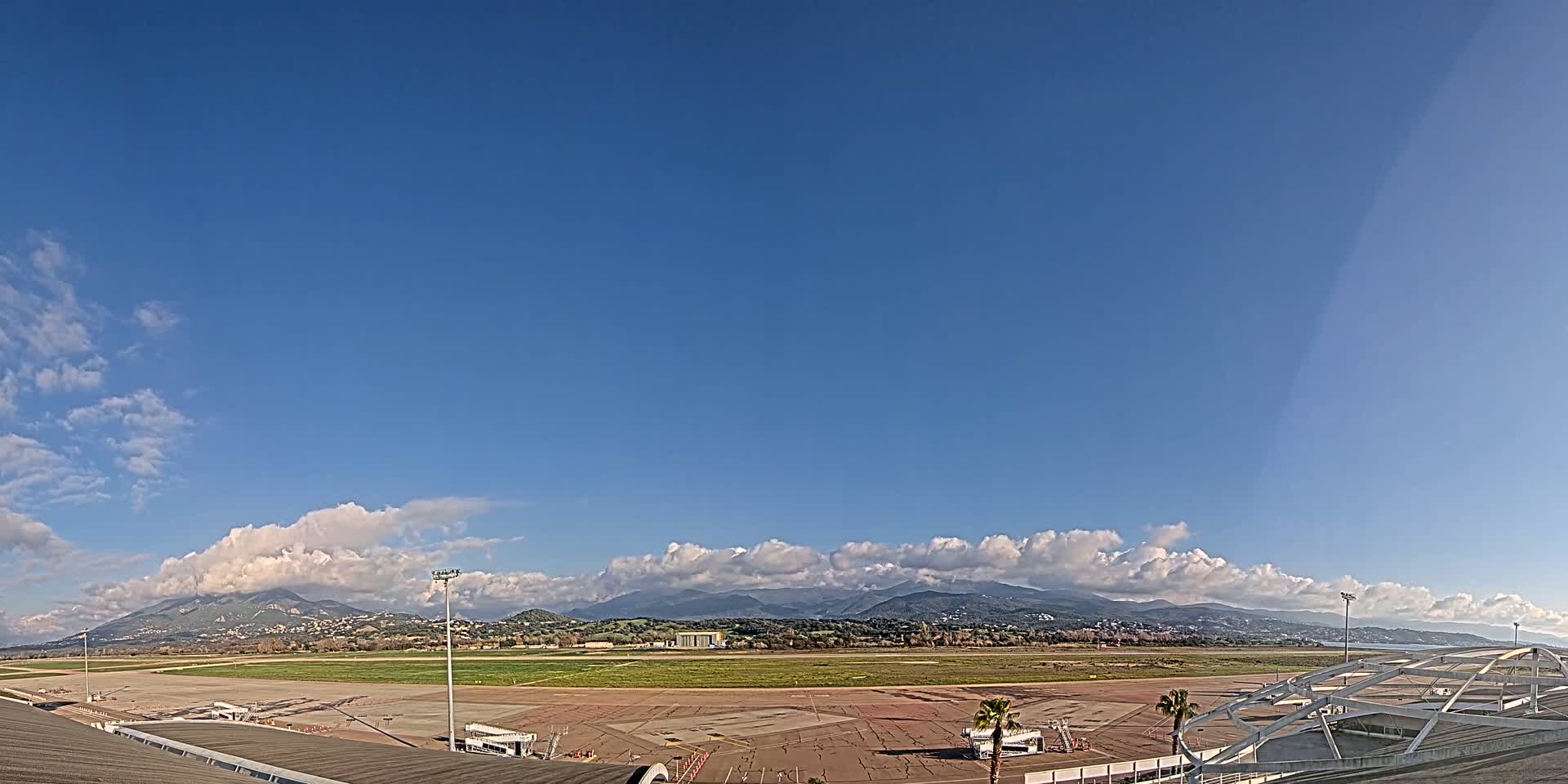 A mostly sunny day with scattered clouds over a large airport tarmac and runway, backed by distant mountains.