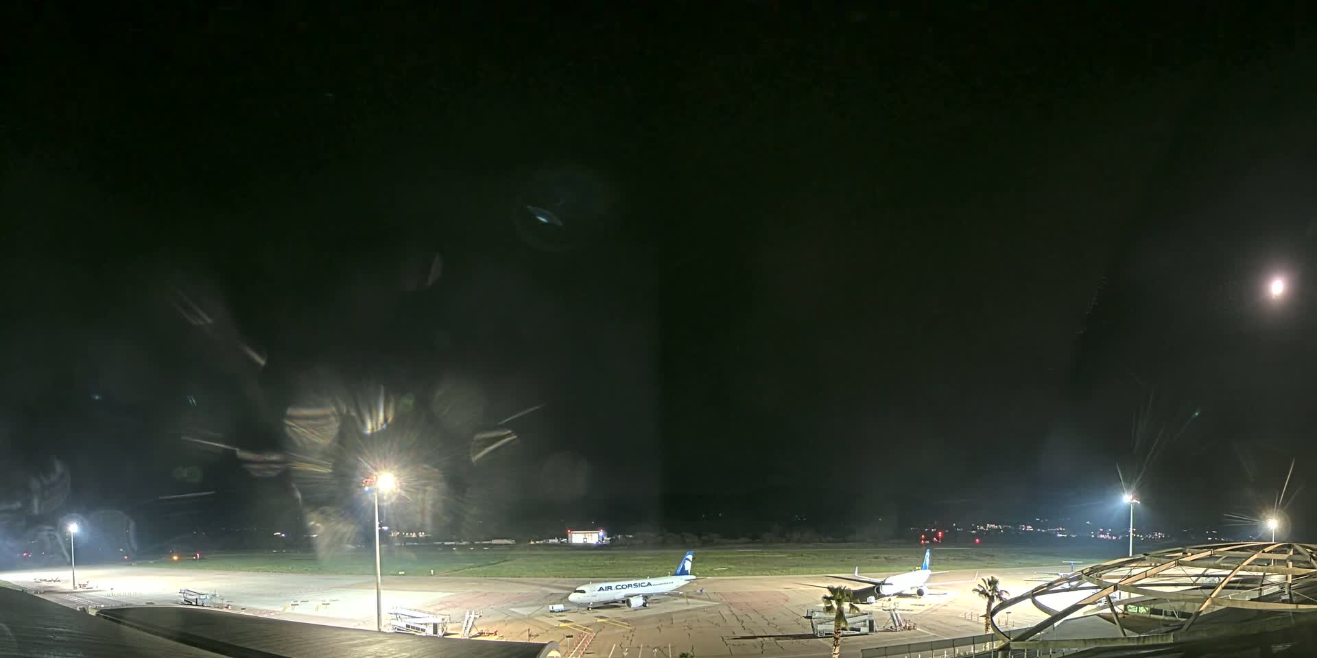 A nighttime view of an airport tarmac with two airplanes parked near a terminal building under a mostly clear sky.