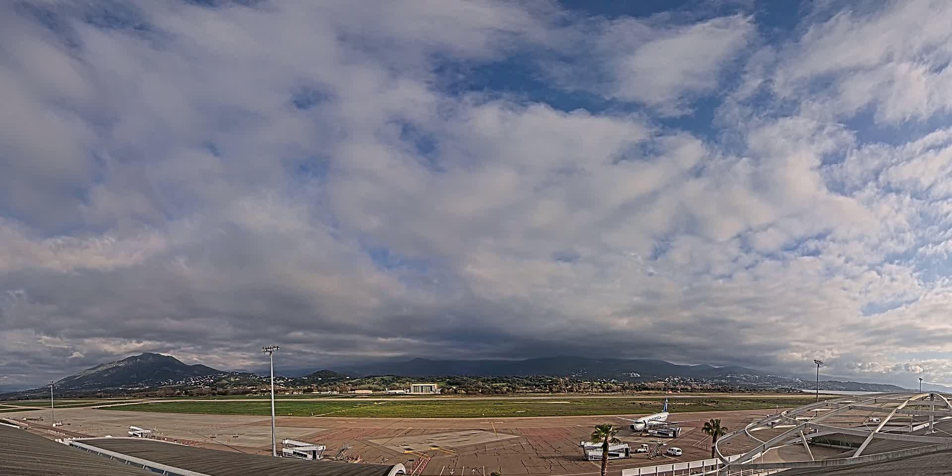 A mostly cloudy sky overlooks an airport tarmac with a few airplanes and surrounding landscape featuring distant mountains.