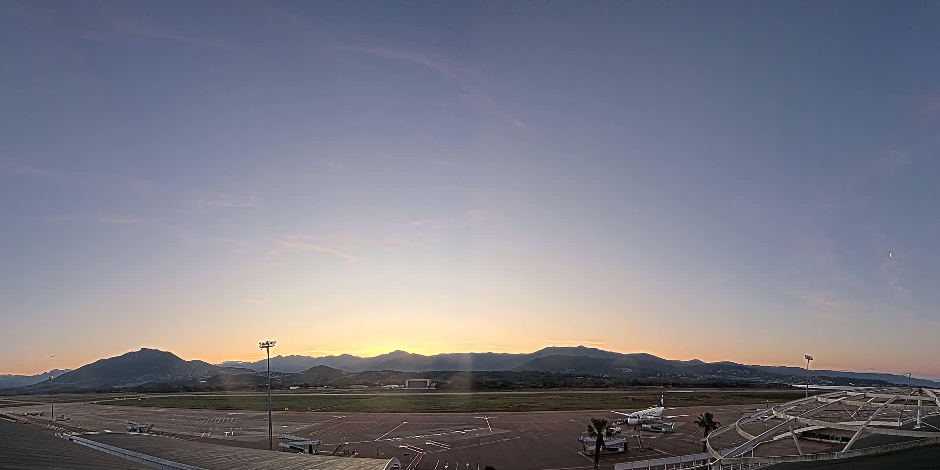 A sunrise illuminates an airport tarmac with a single airplane parked near a terminal building, mountains in the background under a mostly clear sky.