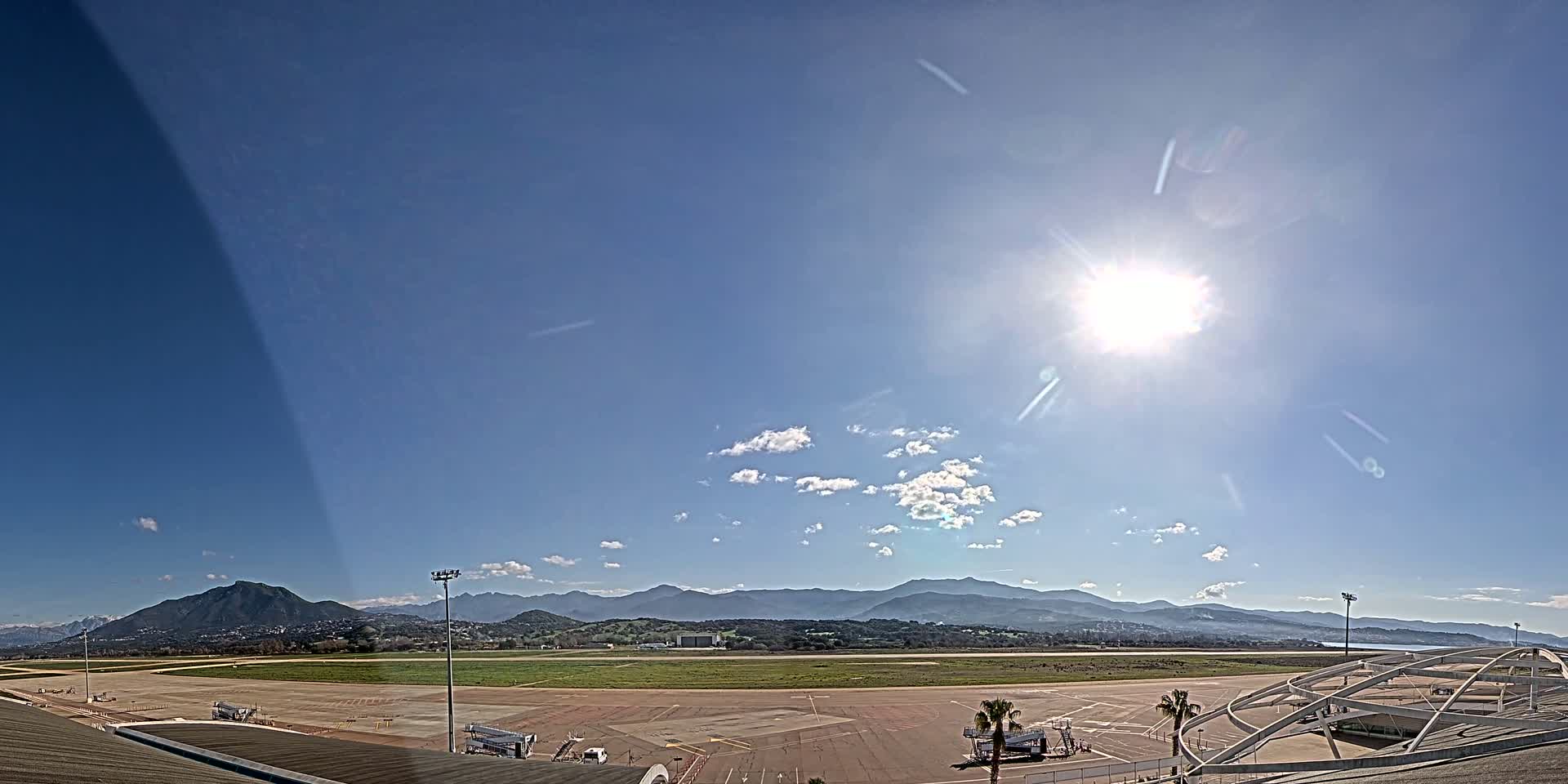 A sunny day reveals a view of an airport tarmac, runways, and distant mountains under a mostly clear blue sky with a few scattered clouds.