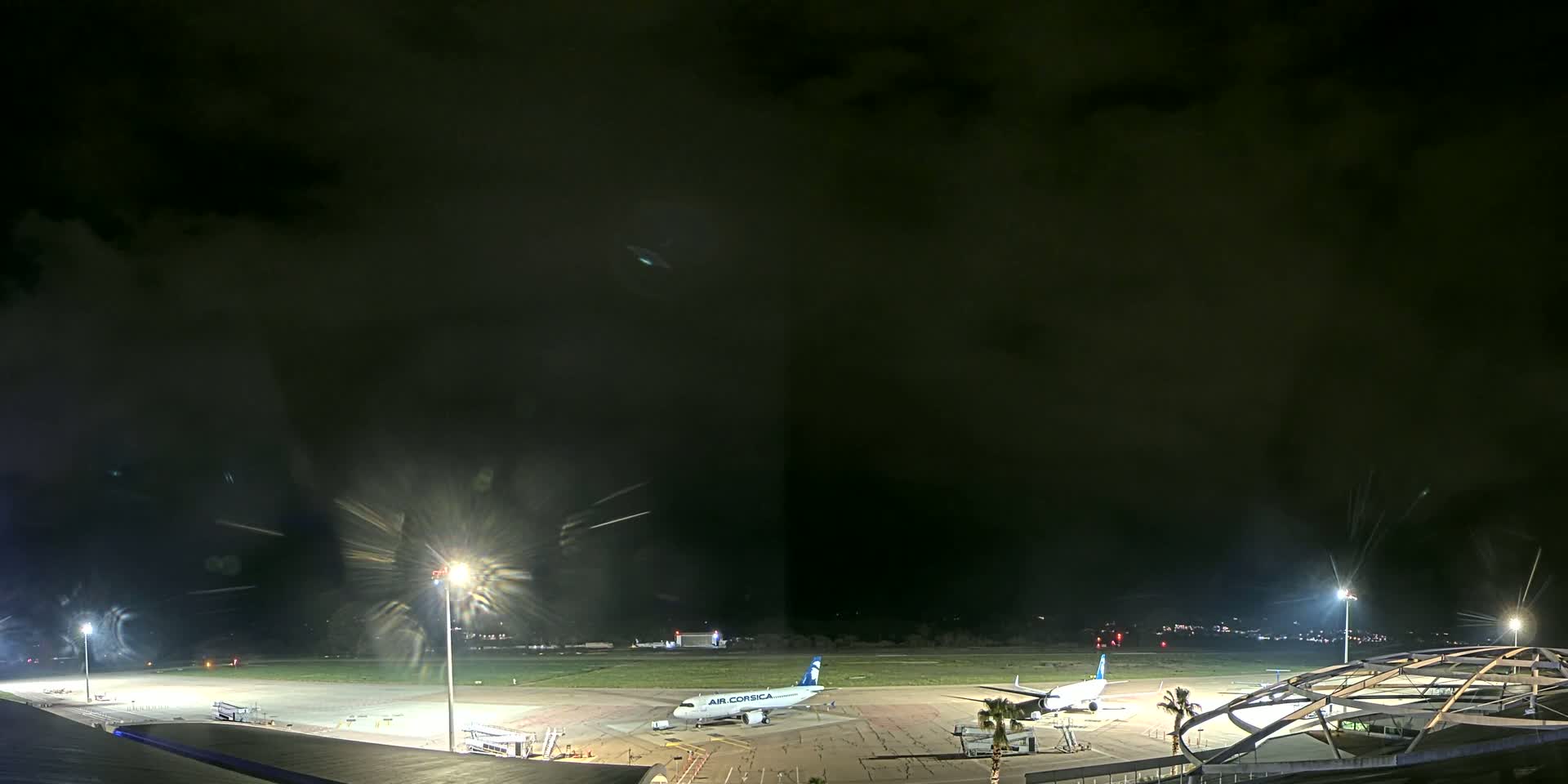 A nighttime view of an airport tarmac with two airplanes parked, under a dark, cloudy sky.
