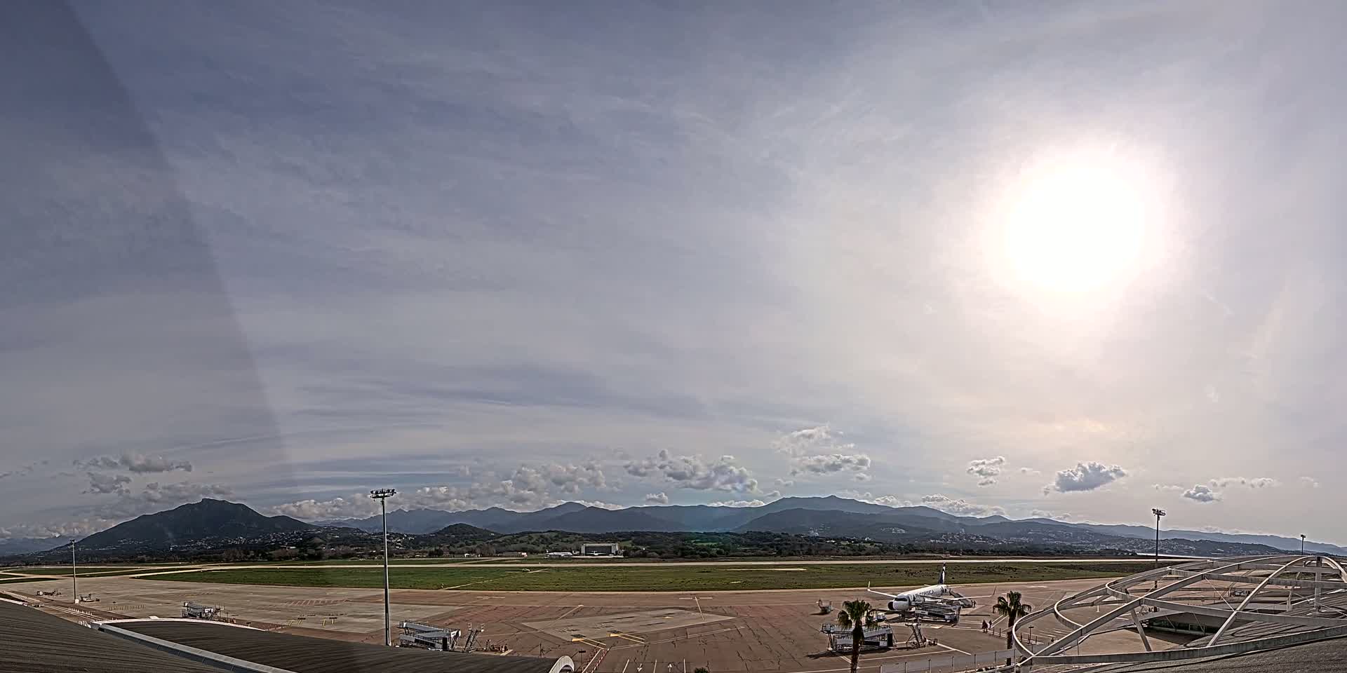 An airport tarmac with a plane parked at a gate, under a mostly sunny sky with scattered clouds and mountains in the distance.