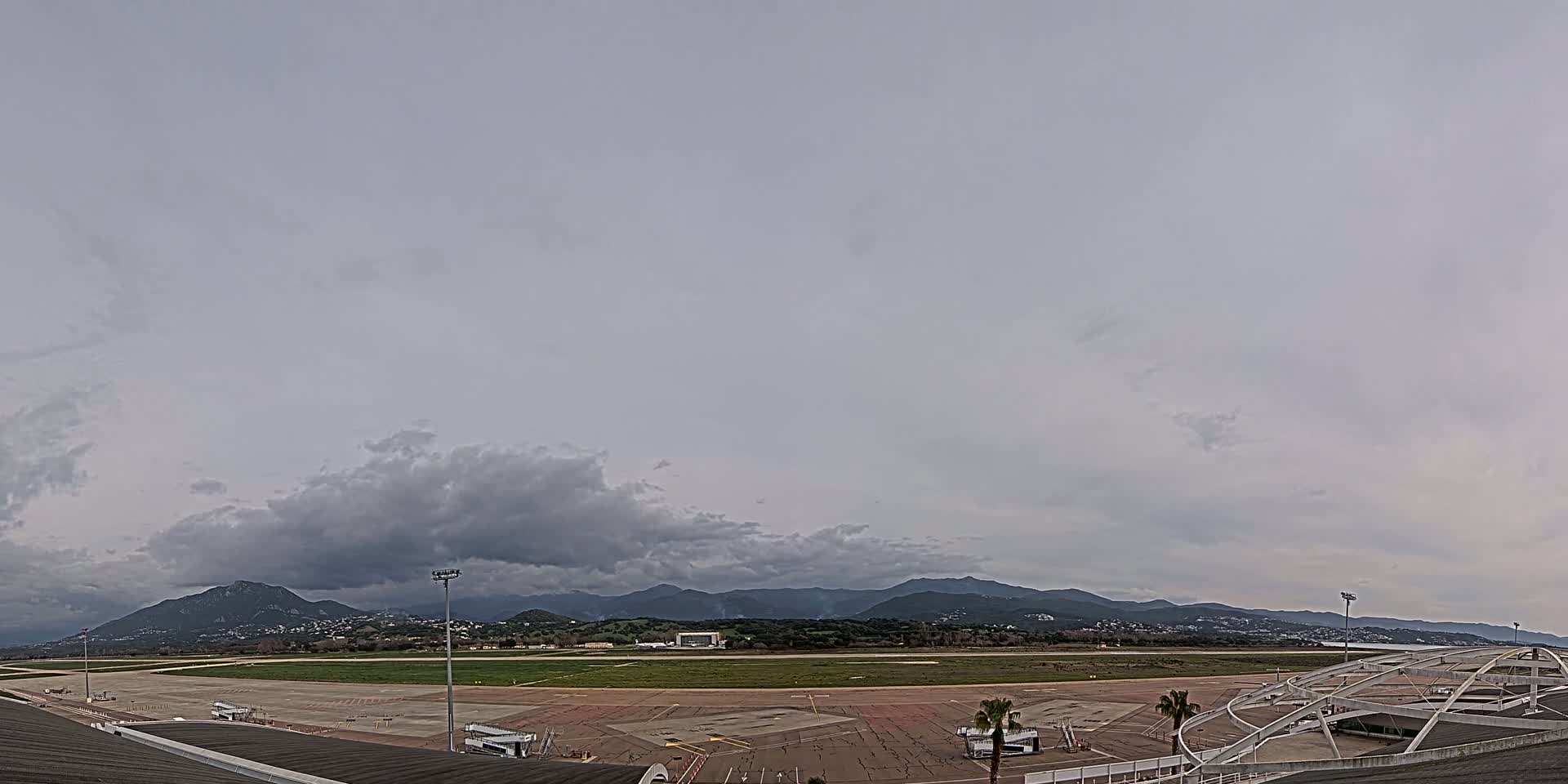An airport tarmac and runway are visible beneath an overcast sky, with mountains in the background.