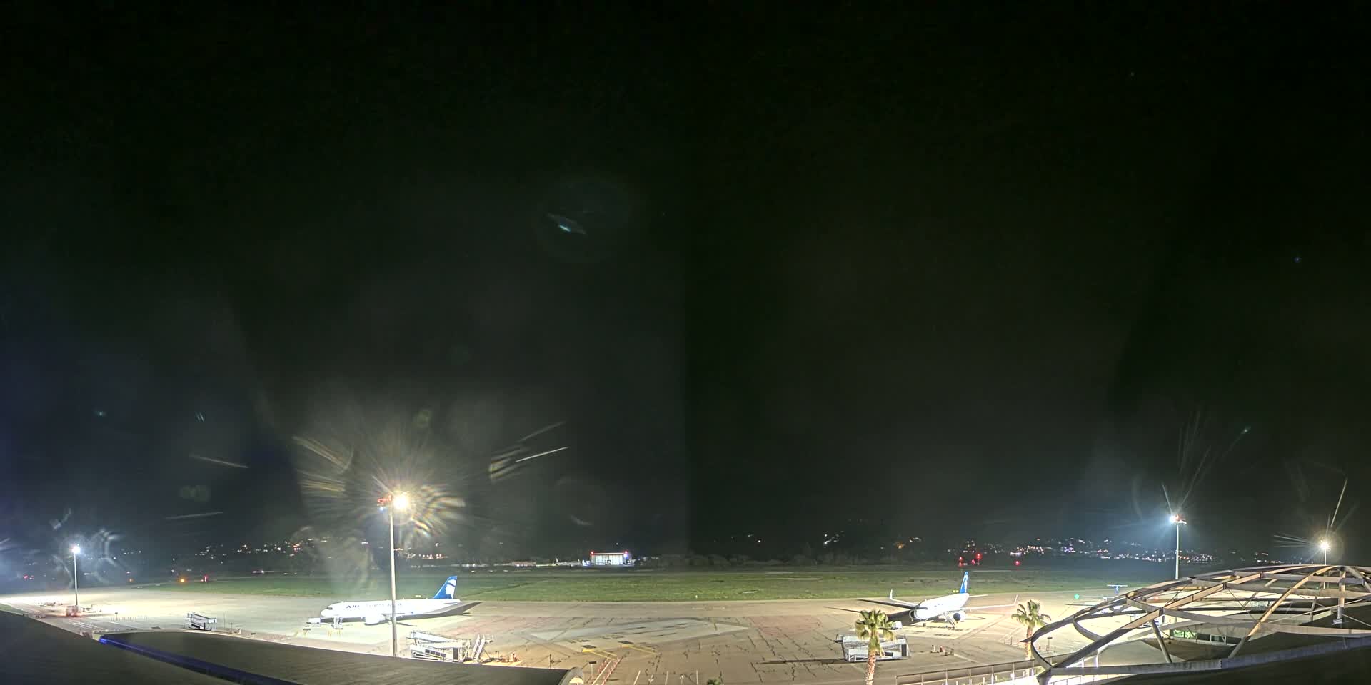 At night, two airplanes are parked on a brightly lit tarmac at an airport.