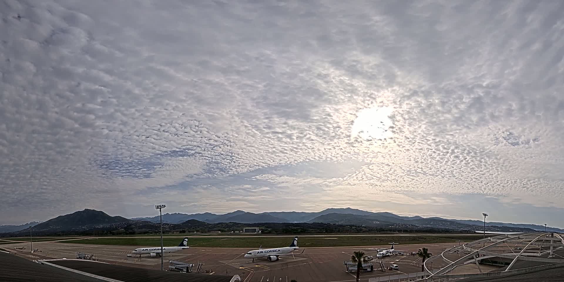 Two airplanes are parked on an airport tarmac under a mostly cloudy sky with the sun visible, backed by distant mountains.