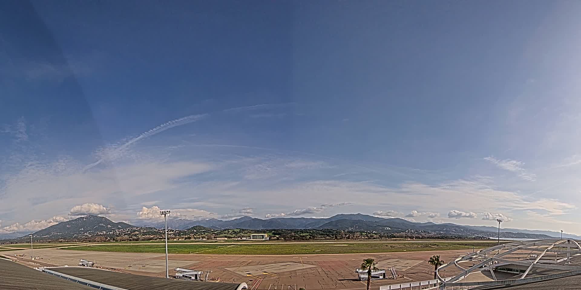 A mostly clear blue sky with some scattered clouds overlooks an airport tarmac and runway, backed by distant mountains.
