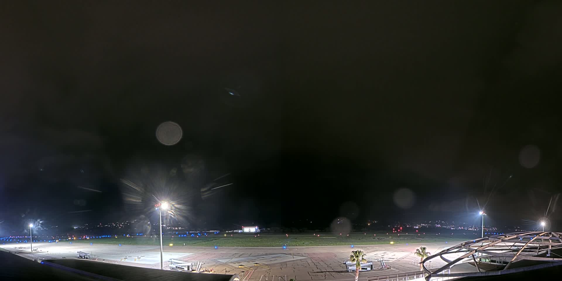 A nighttime view of an airport tarmac with several planes parked and illuminated by bright lights under a dark, possibly stormy sky.