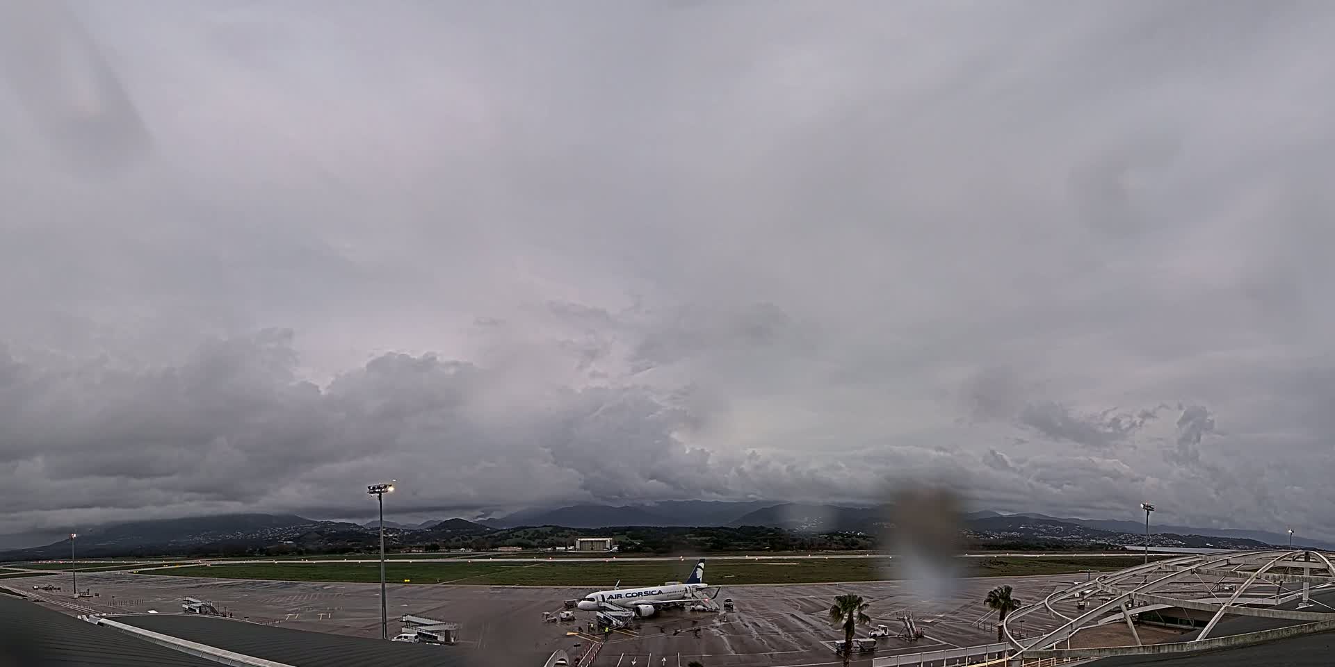 A passenger airplane sits on a tarmac at an airport under a cloudy, overcast sky, with rain apparent on the camera lens.