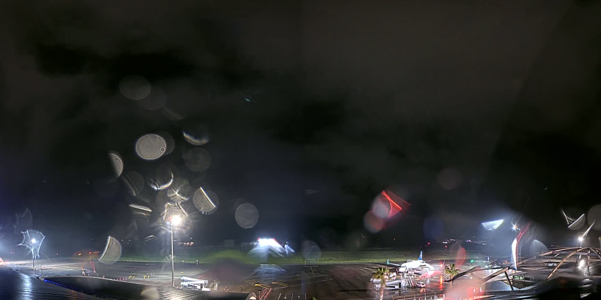 Rain streaks a nighttime view of an airport tarmac with several planes and airport buildings illuminated by lights.