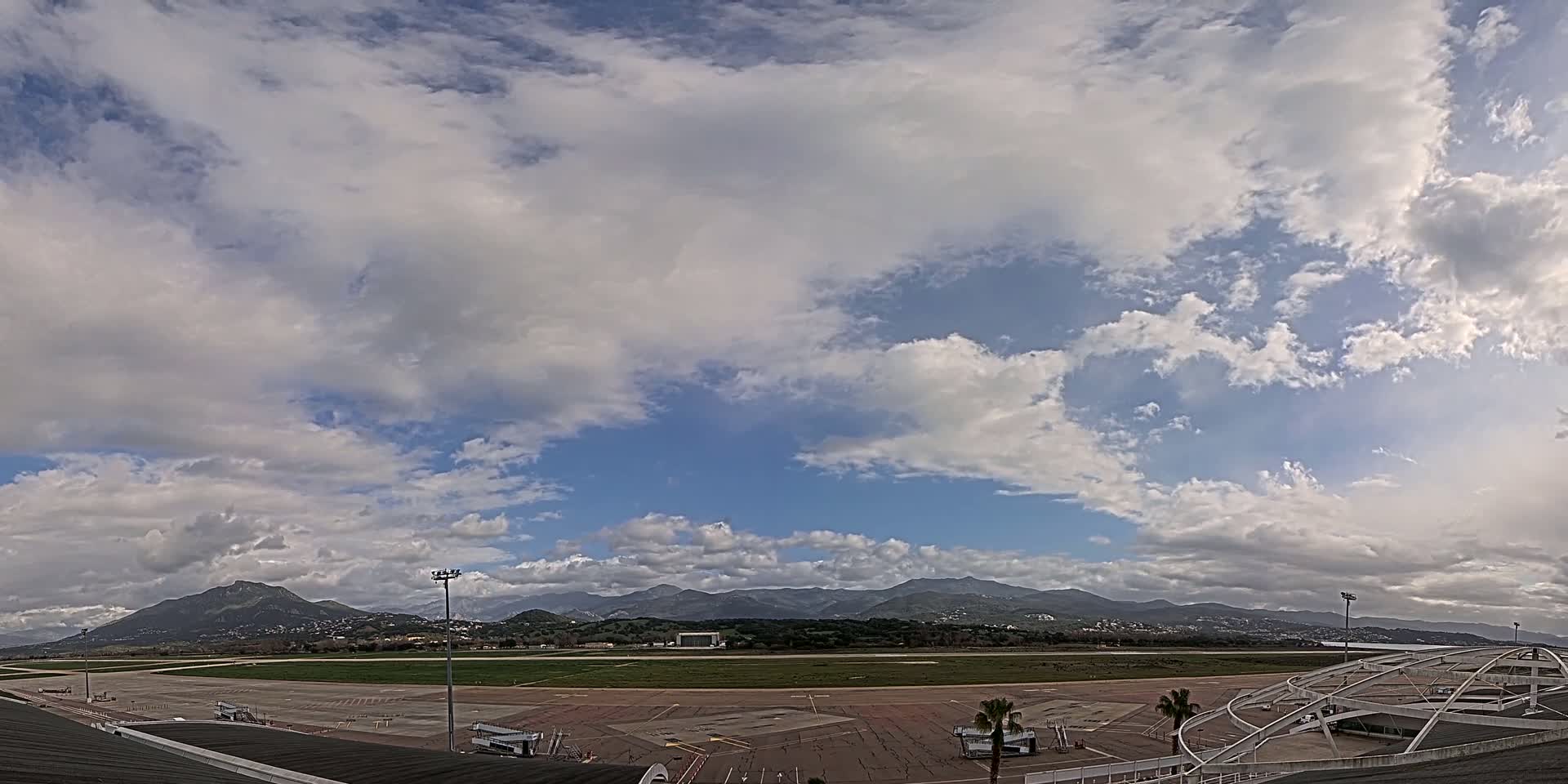 A mostly sunny day with scattered cumulus clouds shows an airport tarmac, runways, and distant mountains.