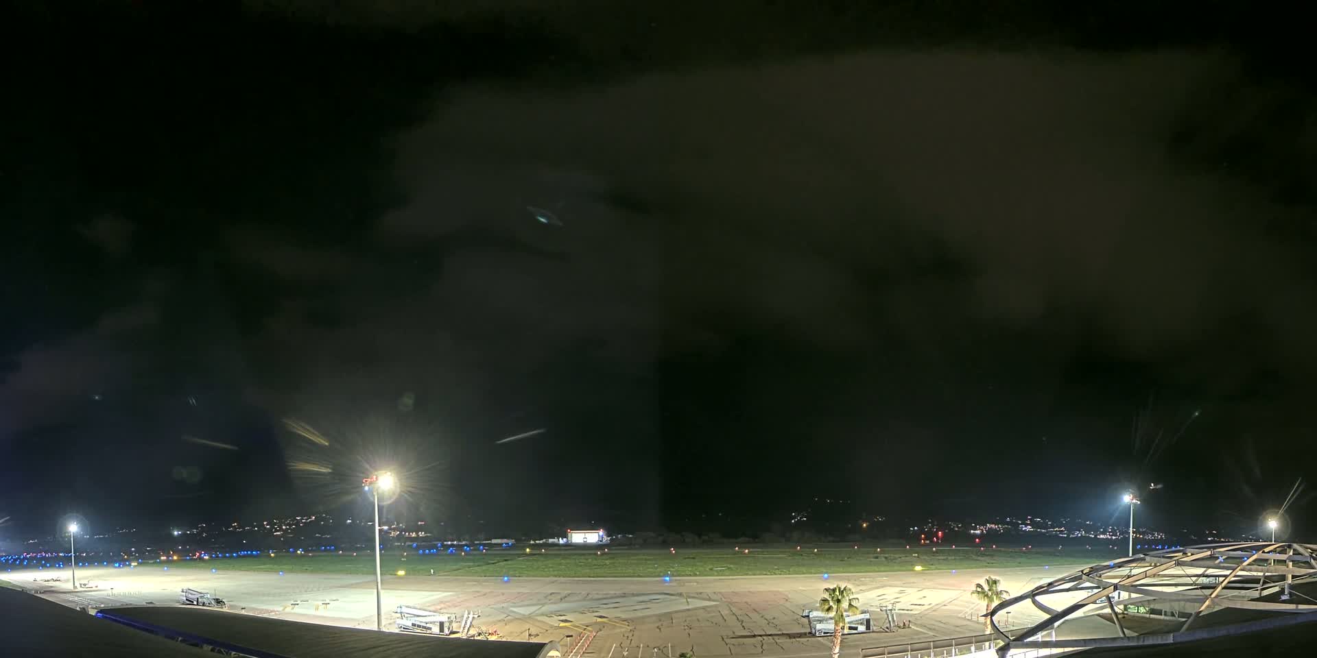 An airport tarmac at night under a dark, cloudy sky is illuminated by tall lights, with some ground vehicles and a building visible.