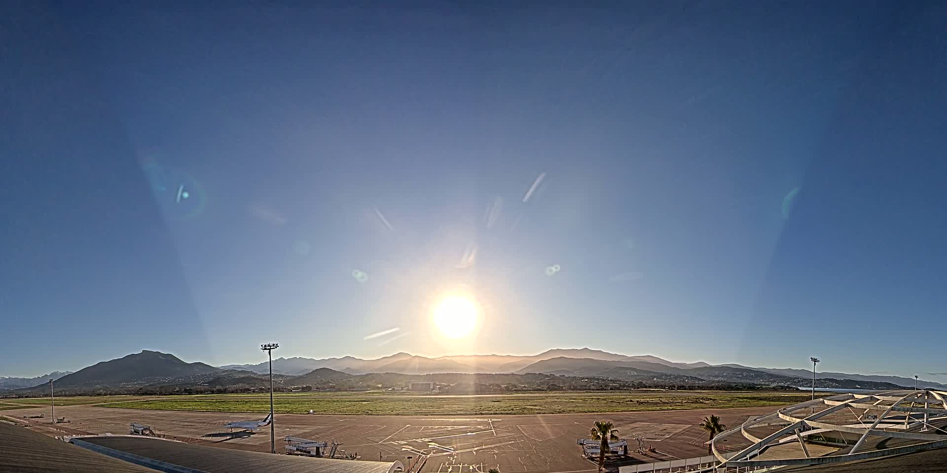 A bright sun shines over a mostly empty airport tarmac with several planes parked, and mountains in the distance under a clear blue sky.