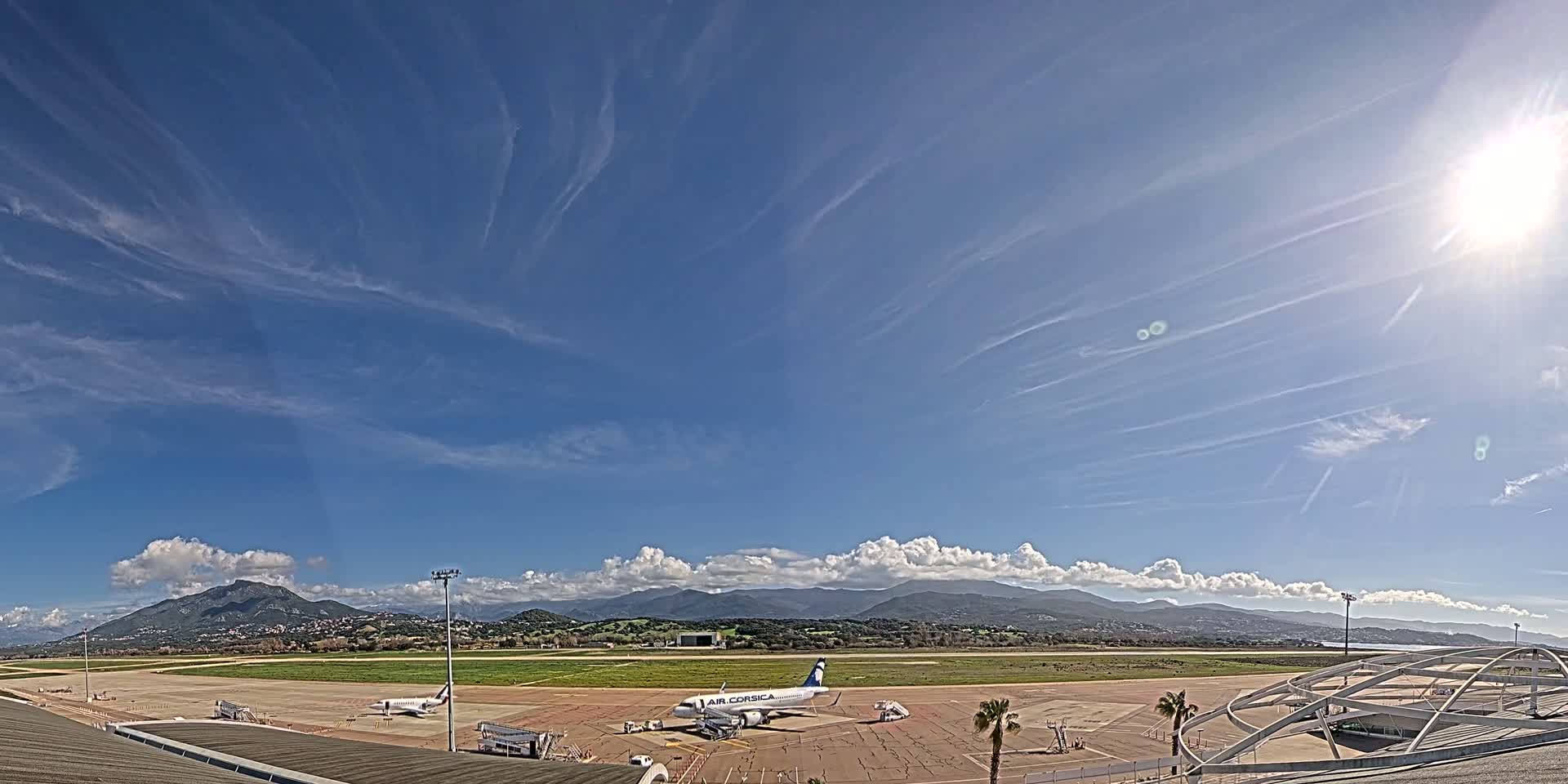 A sunny day at an airport shows several airplanes on the tarmac, with mountains visible in the distance under a mostly clear sky with wispy cirrus clouds.