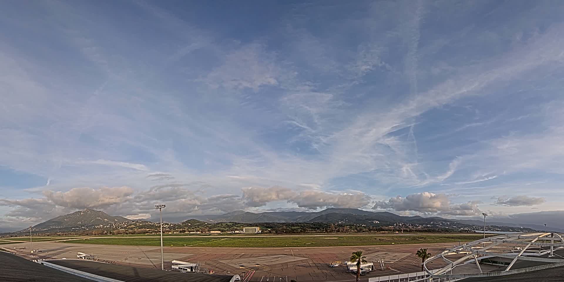 A mostly sunny day with scattered clouds over an airport tarmac and runway, backed by a range of mountains.