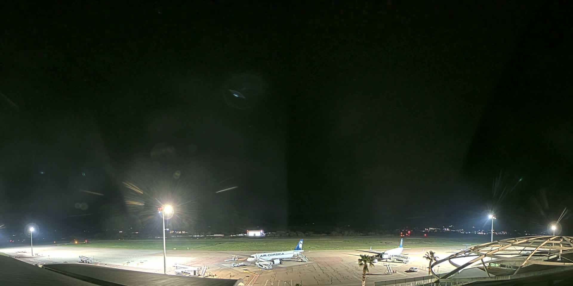 A nighttime view of an airport tarmac with two airplanes parked at gates, illuminated by bright lights under a clear, dark sky.
