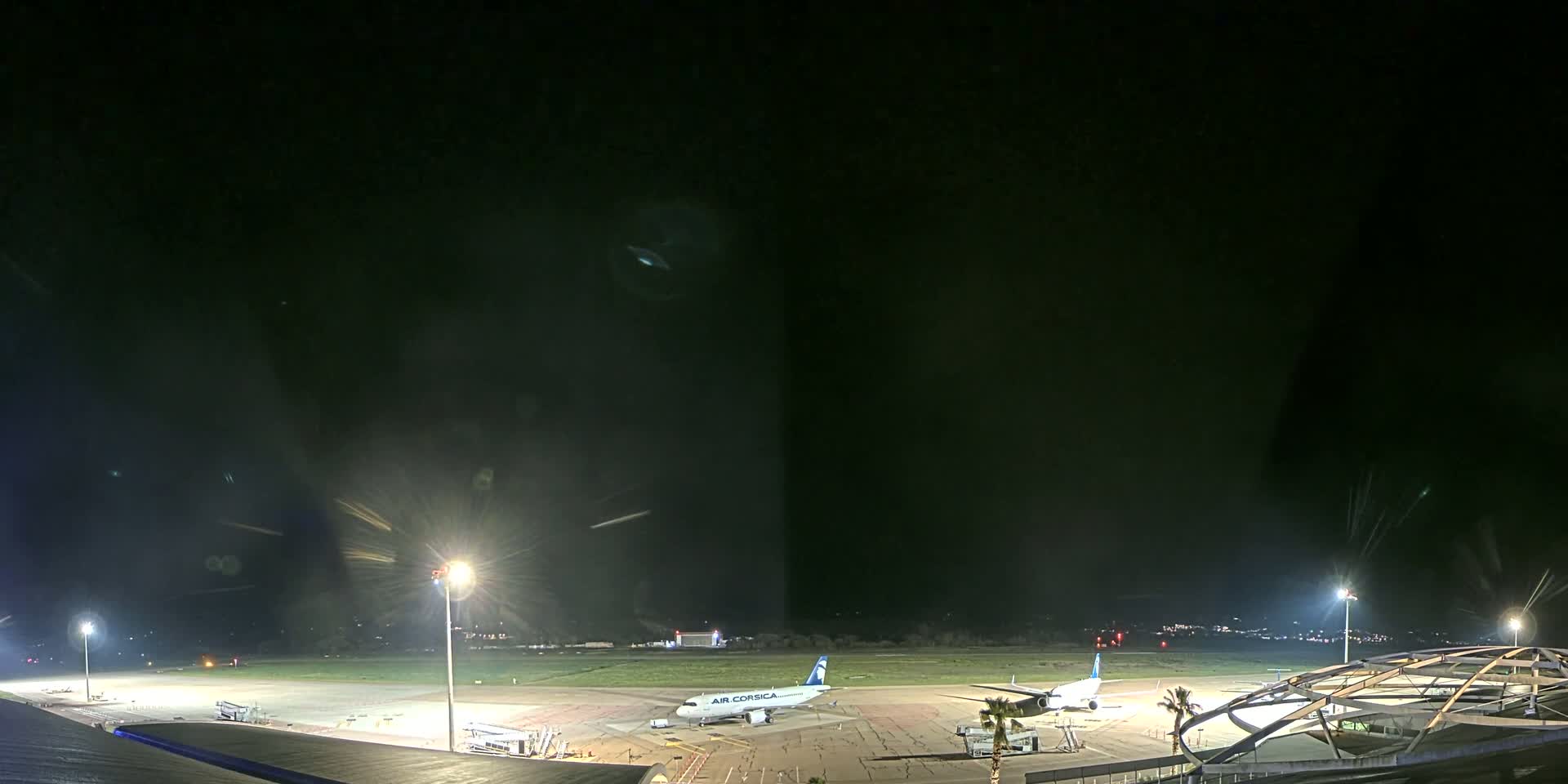 At night, two airplanes are parked on an airport tarmac illuminated by bright lights, under a dark, clear sky.