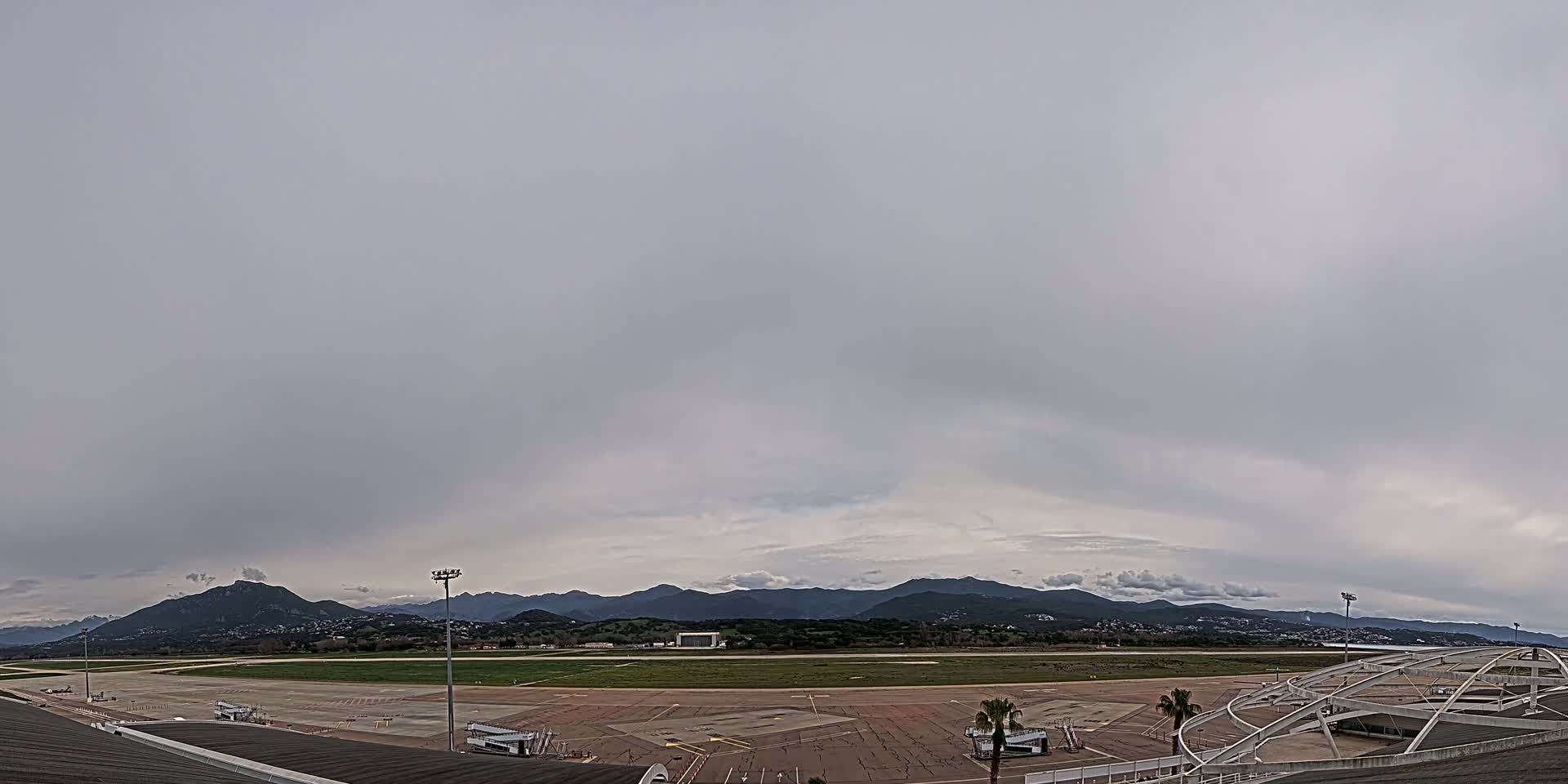 An airport tarmac and runway are visible beneath an overcast sky, with mountains in the distance.
