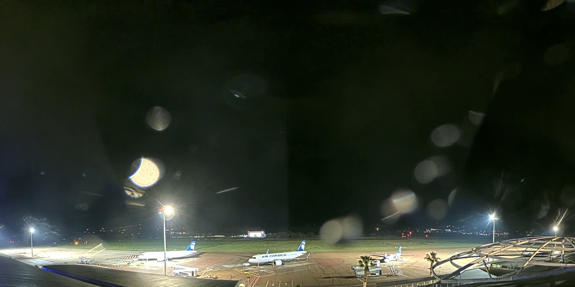 At night, under a dark and rainy sky, two airplanes are parked on a tarmac at an airport, surrounded by lights and airport structures.