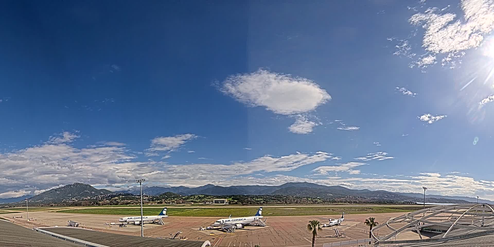 Two airplanes sit on a tarmac at an airport under a mostly sunny sky with scattered clouds and distant mountains visible in the background.