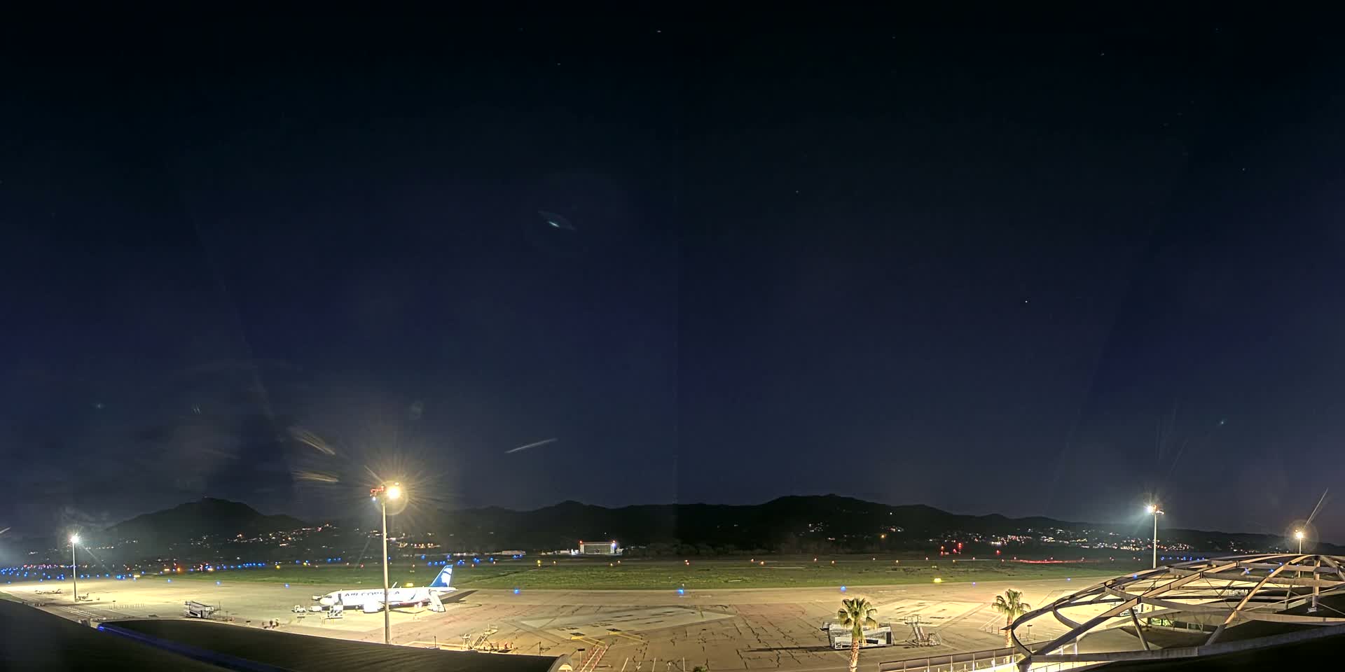 A nighttime view of an airport tarmac with a plane parked at a gate, illuminated by bright lights against a dark sky with some stars visible, and mountains in the background.