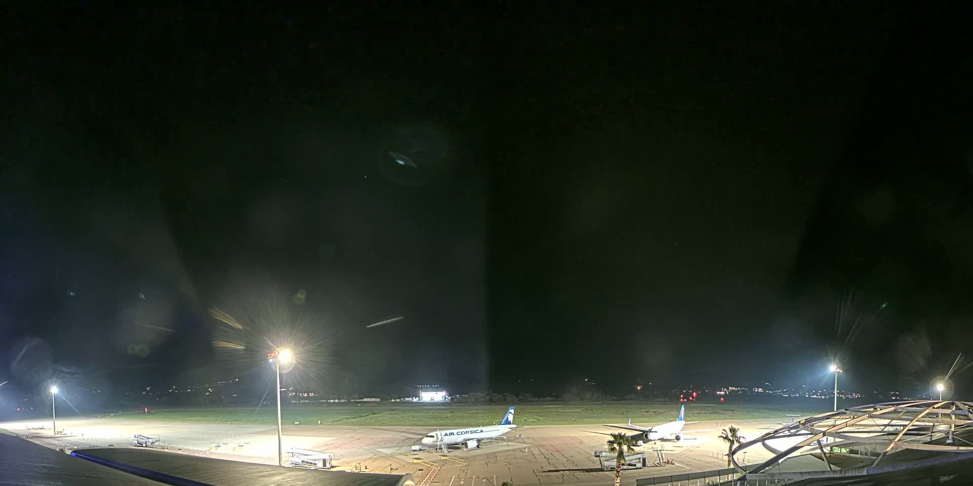A nighttime view of an airport tarmac with two airplanes parked, illuminated by bright lights under a dark sky.