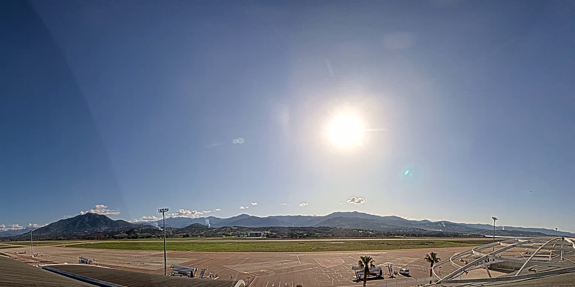 A sunny day at an airport with a runway, parking lot, and mountains in the distance.