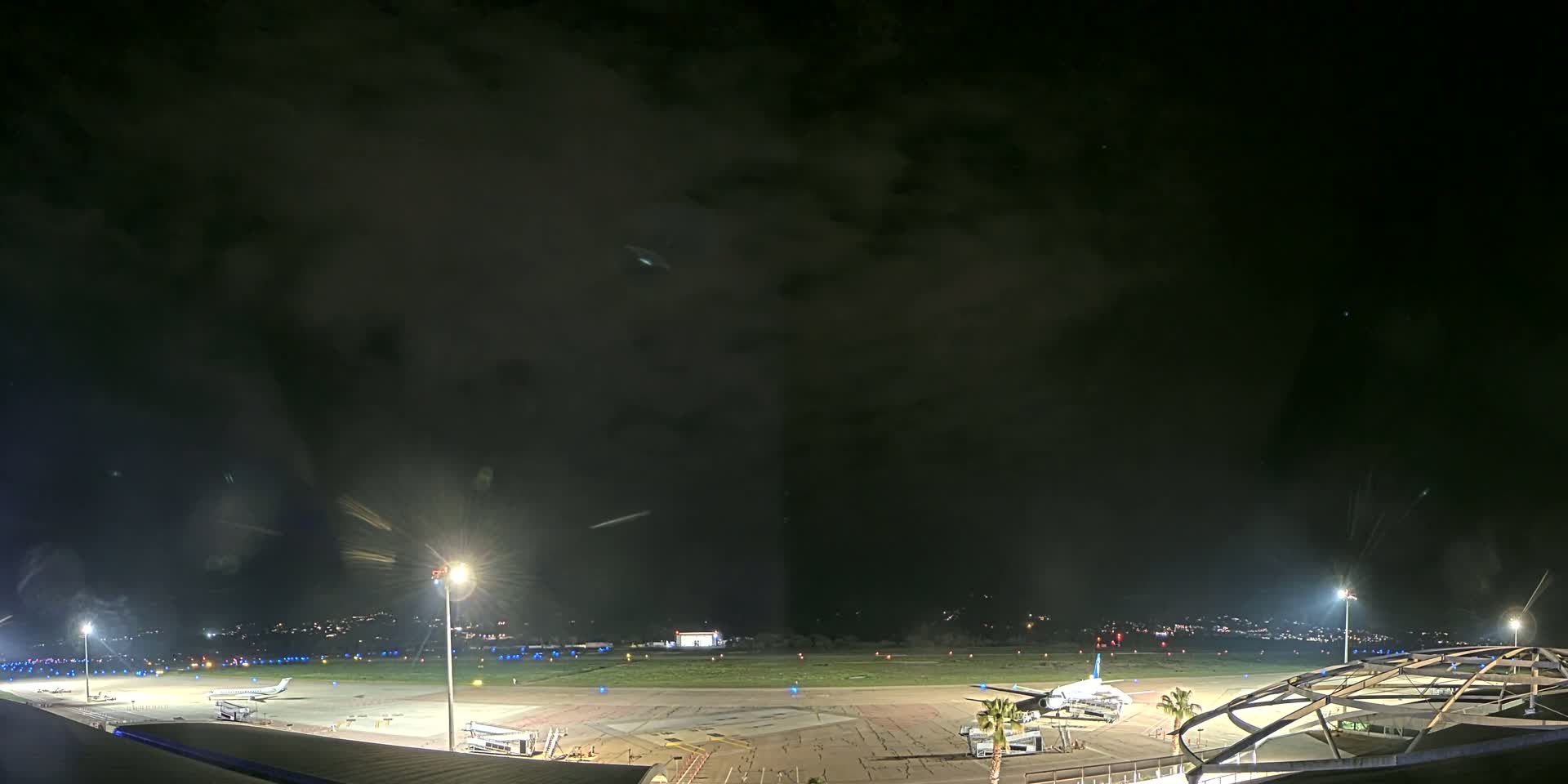 A nighttime view of an airport tarmac with several airplanes parked, illuminated by bright lights under a dark, cloudy sky.
