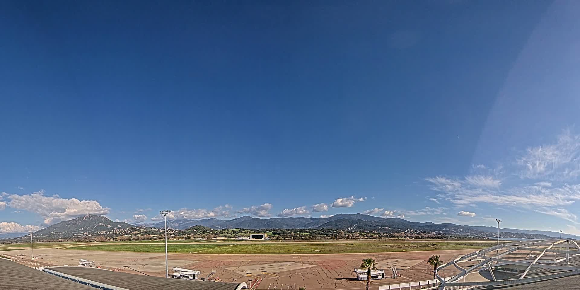 An airport tarmac and runway extend toward a range of mountains under a mostly clear blue sky with some scattered clouds.
