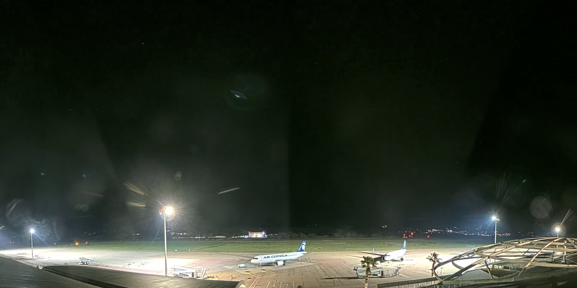 A nighttime view of an airport tarmac with two airplanes parked near airport buildings under bright lights.
