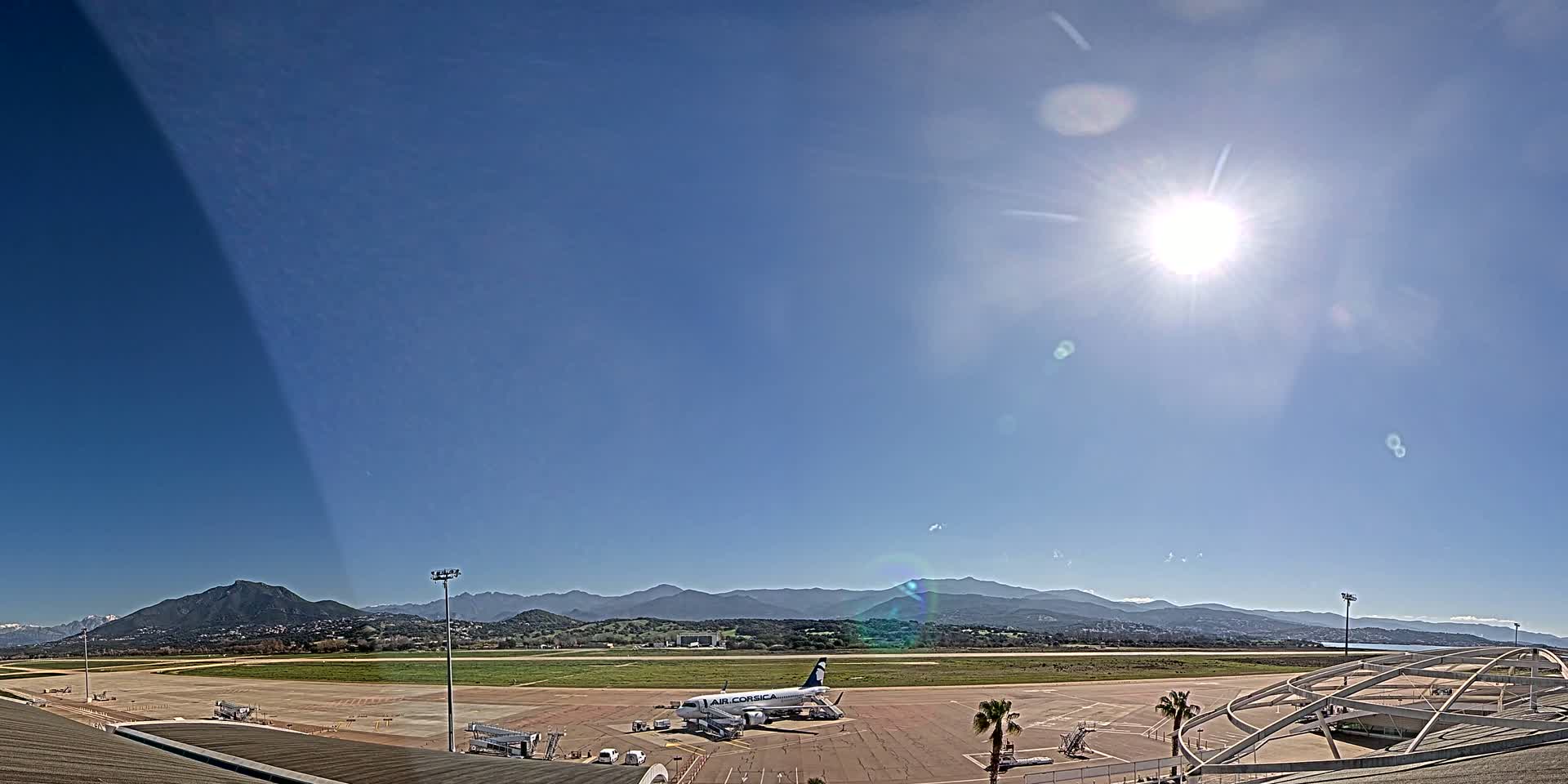 A bright, sunny day at an airport with a single airplane parked on the tarmac, mountains visible in the distance.