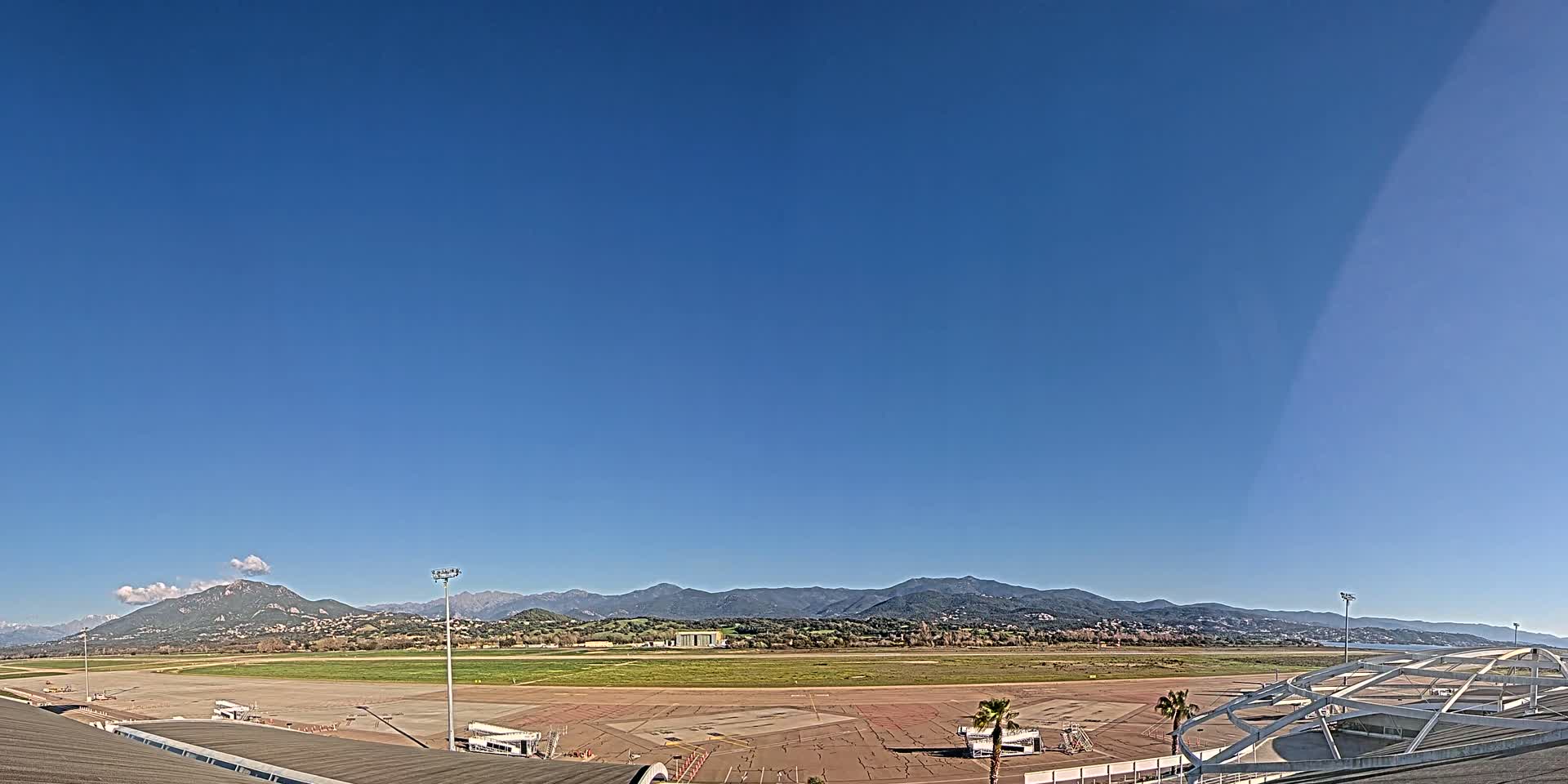 An airport tarmac and runway are situated before a mountain range under a clear blue sky.
