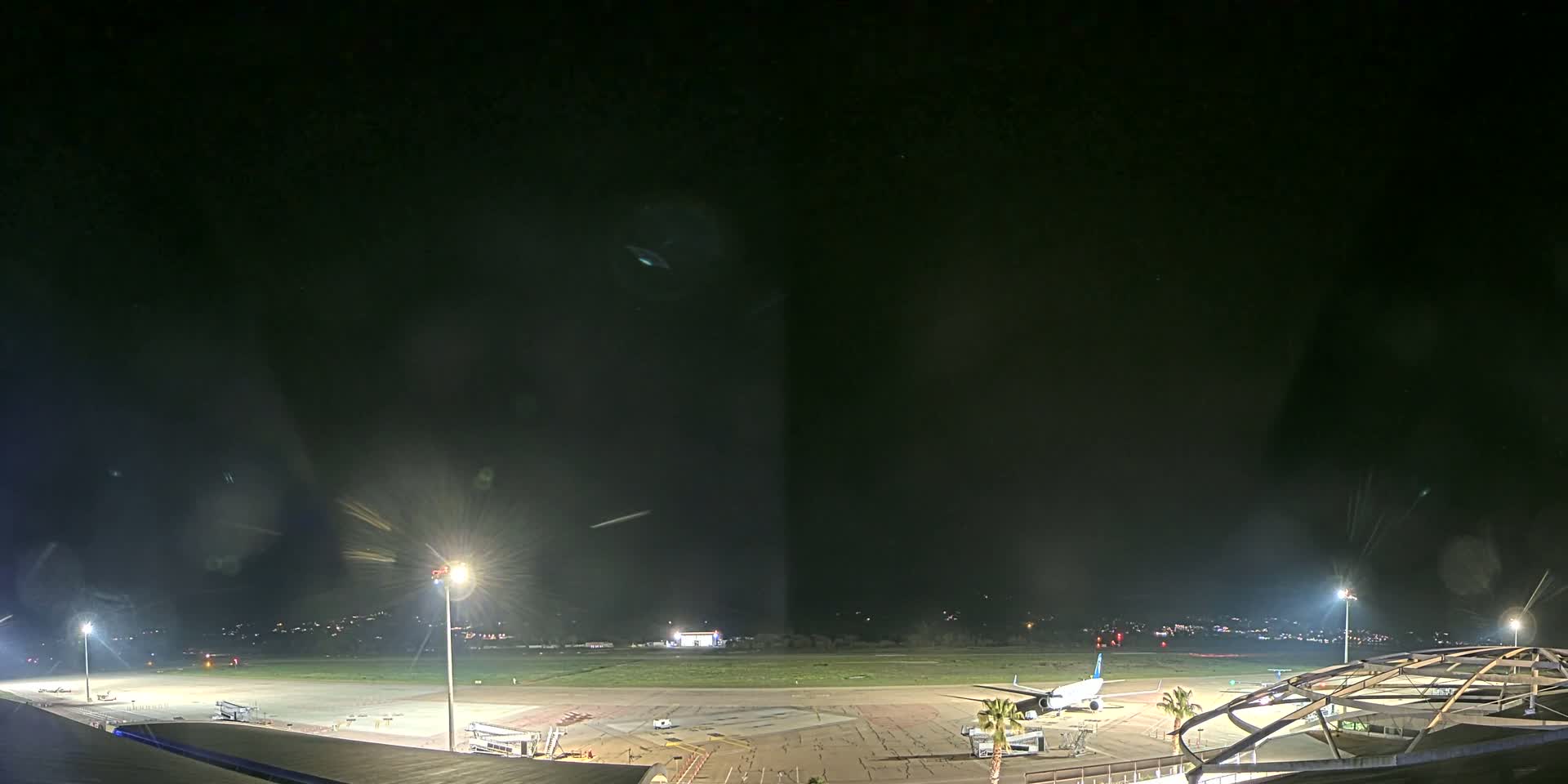 A nighttime view of an airport tarmac with a plane parked near a terminal building under a clear, dark sky.
