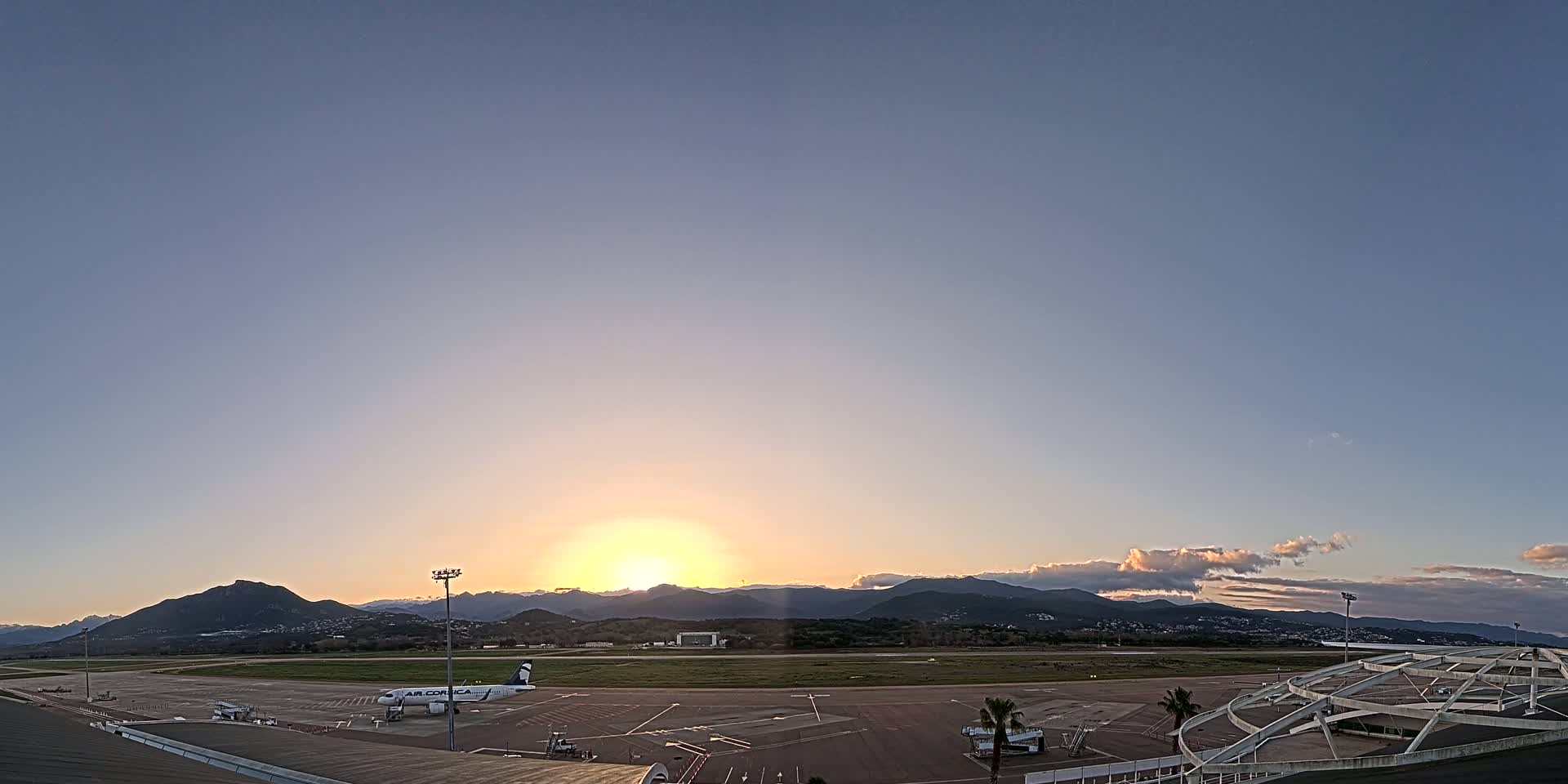 A sunrise casts a warm glow over an airport tarmac with a single airplane parked and mountains in the distance under a mostly clear sky.