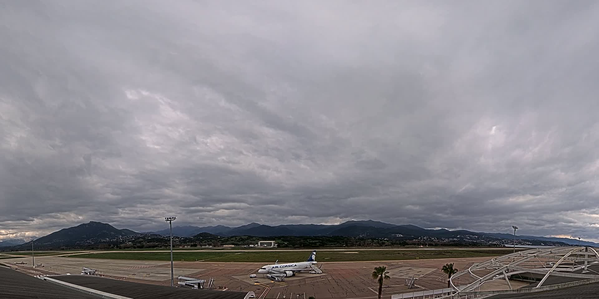 A large passenger airplane sits on an airport tarmac under a heavily overcast sky, with mountains visible in the distance.
