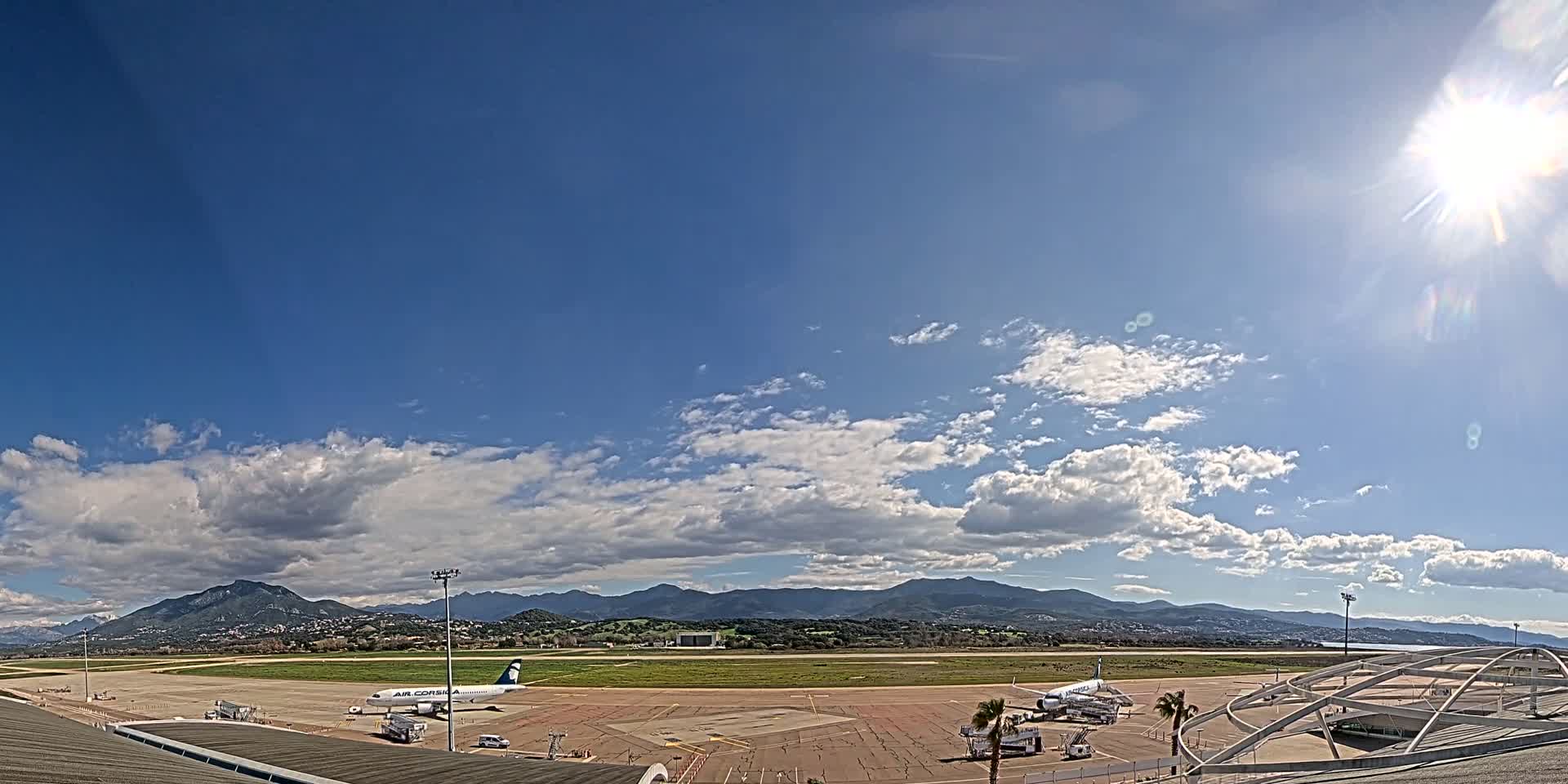 A sunny day with partly cloudy skies over an airport tarmac, showing several airplanes and surrounding mountainous landscape.