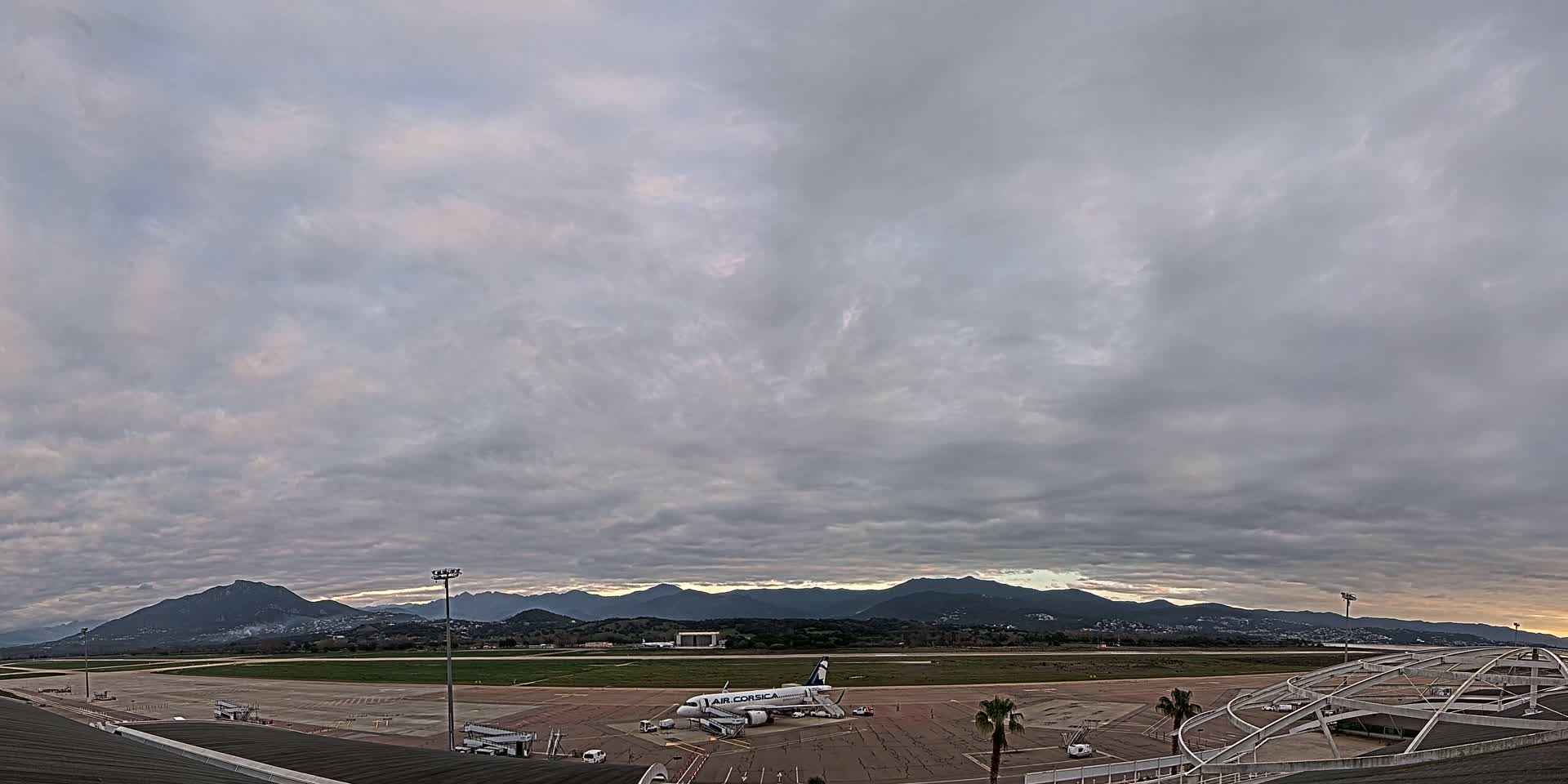 An airplane sits on an airport tarmac under a mostly cloudy sky, with mountains visible in the distance.